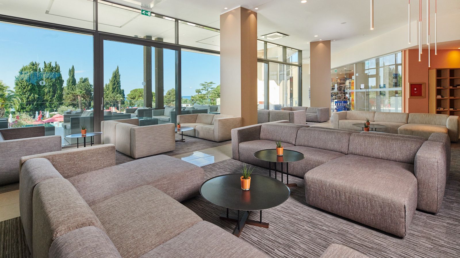 Moderne Lounge mit bequemen Sofas, Tischen und Blick ins Freie auf Bäume und einen blauen Himmel.