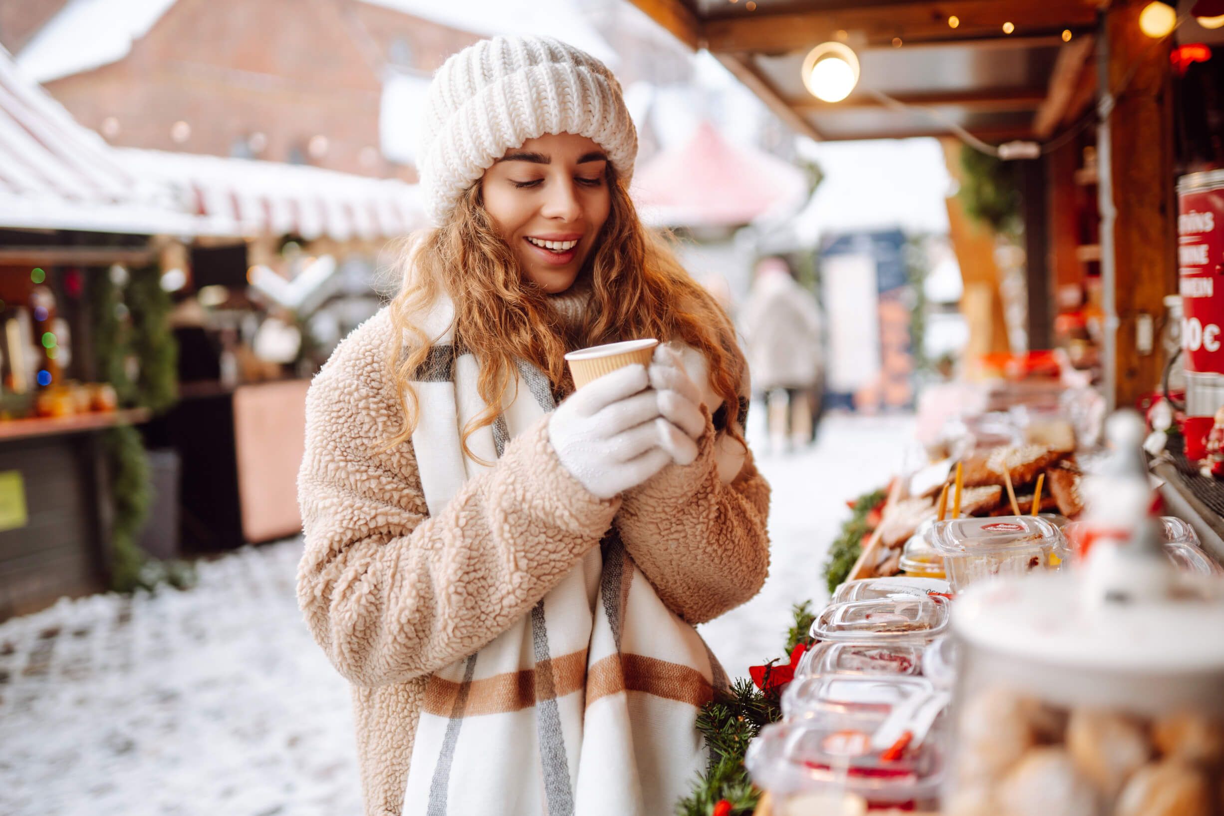 Eine fröhliche Frau mit Mütze genießt ein Getränk auf einem Weihnachtsmarkt im Schnee.