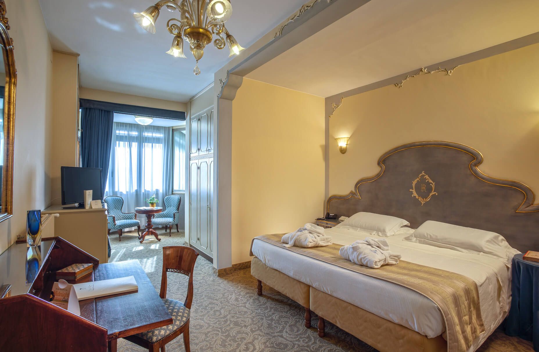 Ein gemütliches Hotelzimmer mit Doppelbett, Schreibtisch und Sitzecke vor großen Fenstern.