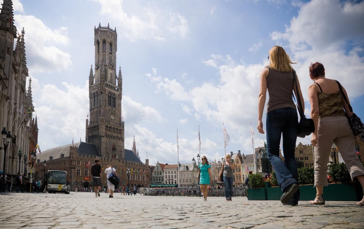 Menschen schlendern durch einen Platz mit einem hohen Glockenturm und historischen Gebäuden unter einem blauen Himmel.