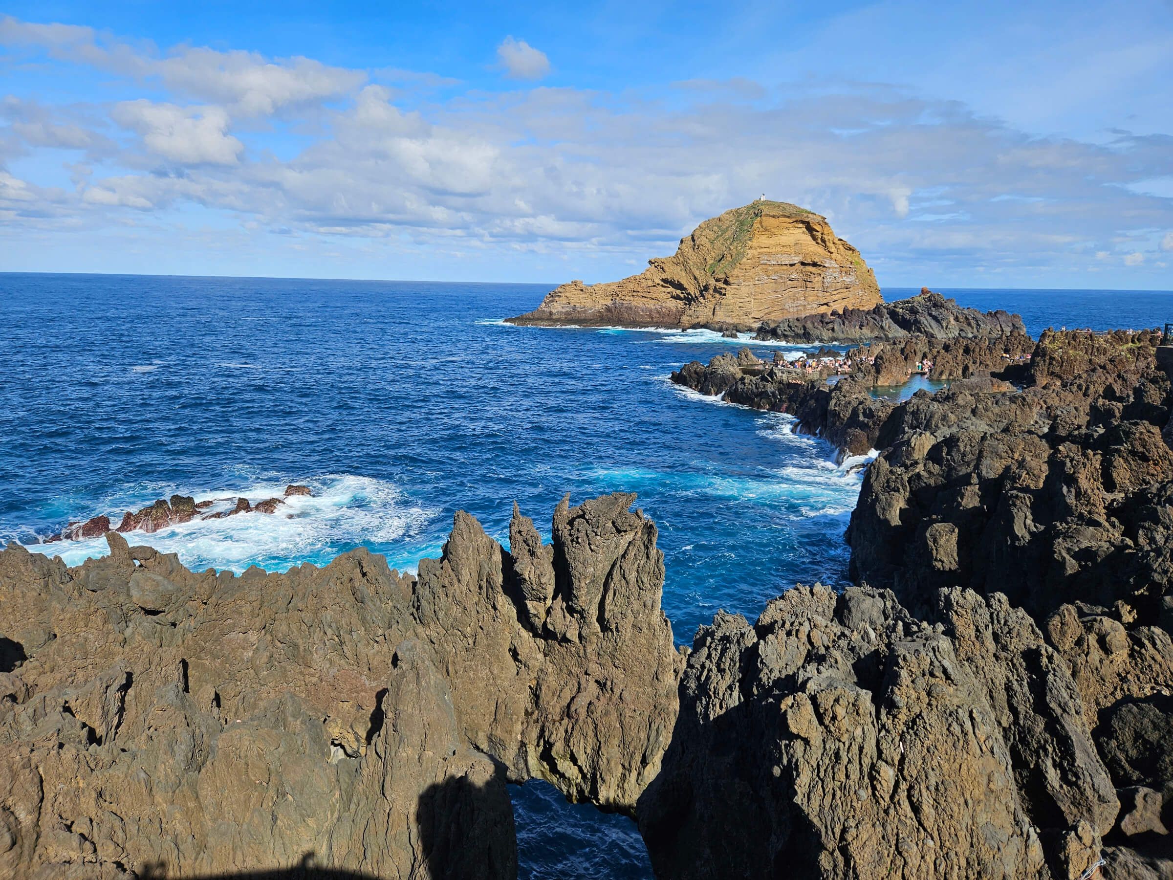 Ein Felsenküstenbild mit blauen Wellen, schroffen Klippen und einer kleinen Insel im Hintergrund.