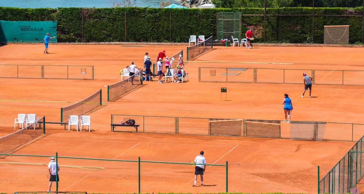 Tennisplätze mit Spielern und Zuschauern, umgeben von grünem Sichtschutz und Blick auf Wasser. sonniger Tag.