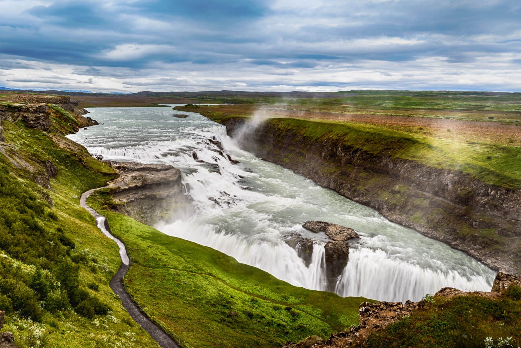 Eine beeindruckende Landschaft mit einem mächtigen Wasserfall, umgeben von grünen Wiesen und dramatischen Wolken.
