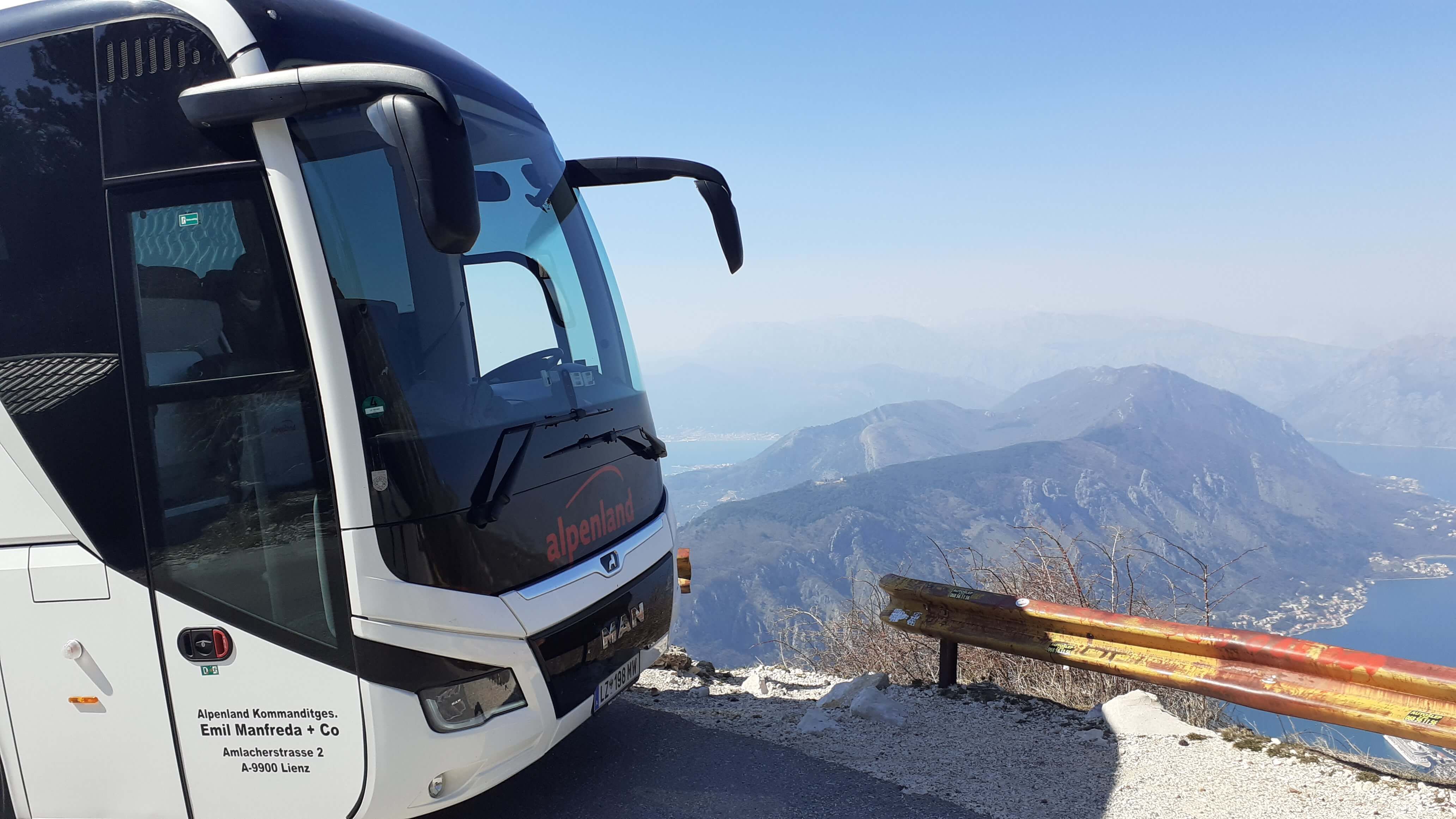 Ein Bus steht an einer Bergstraße mit Aussicht auf Berge und einen See im Hintergrund. Klarer Himmel.