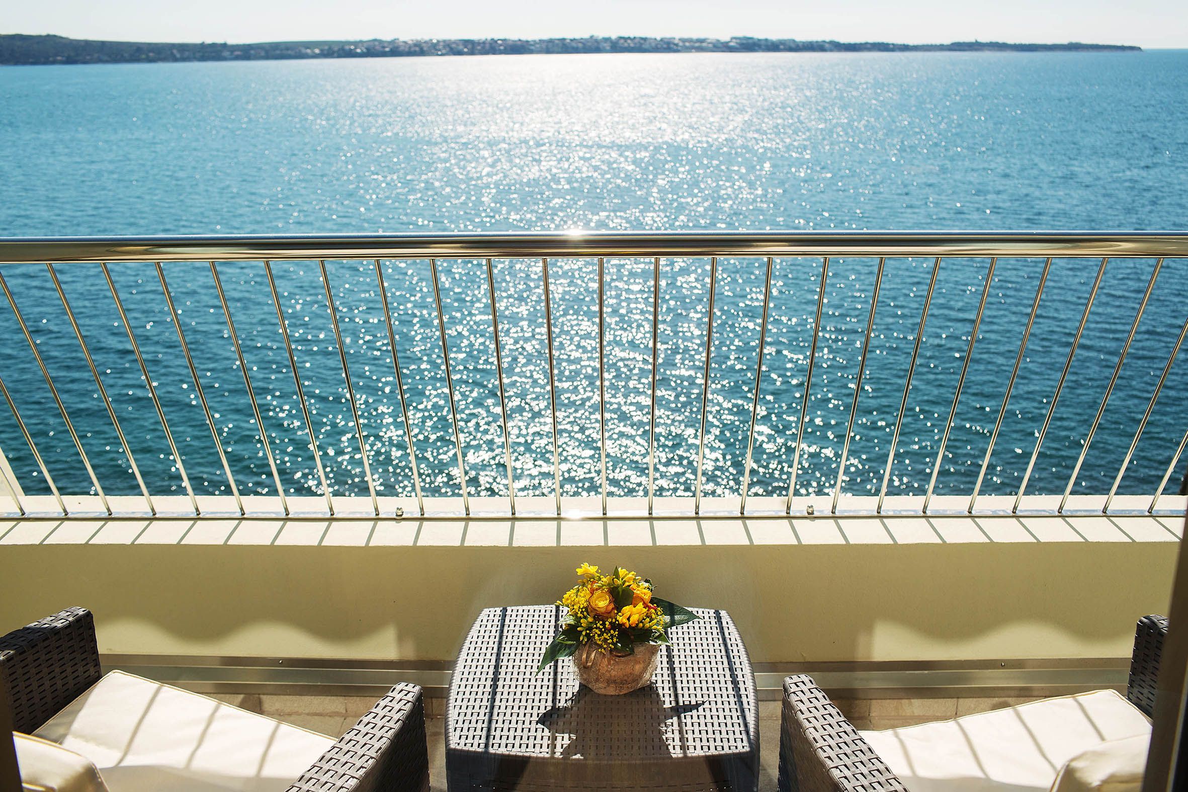 Balkon mit Aussicht auf glitzerndes Wasser und einem Blumenstrauß auf dem Tisch. Entspanntes Urlaubsambiente.