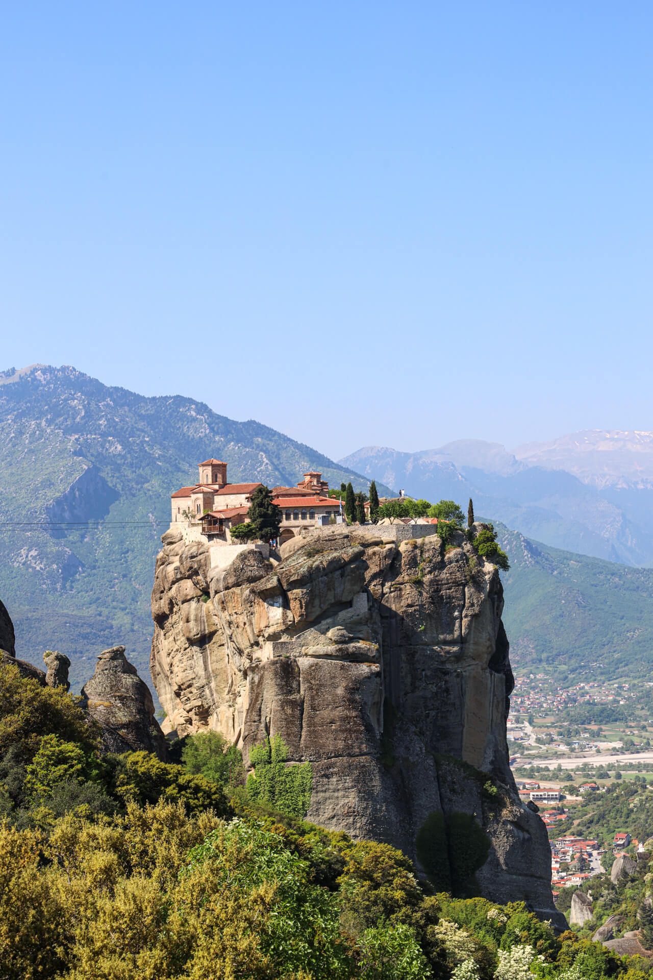 Ein Kloster thront malerisch auf einem steilen Felsen, umgeben von Bergen und grünem Terrain.