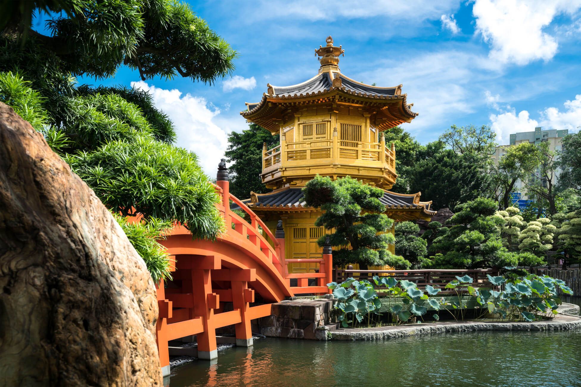 Eine malerische Landschaft mit einem goldenen Pavillon, einer roten Brücke und üppiger Vegetation am Wasser.