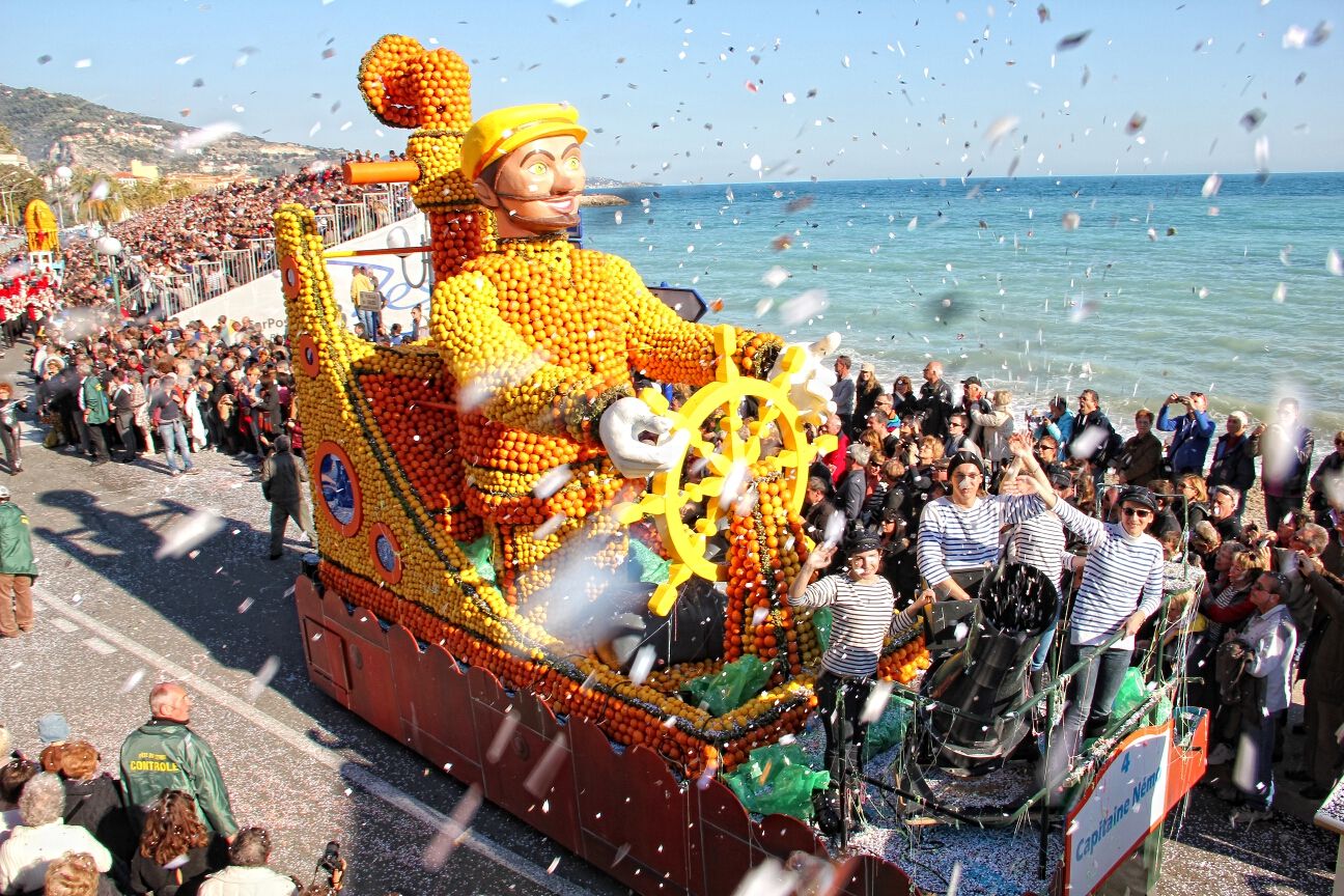 Ein Wagen aus Orangen mit einem Steuermann fährt an einer Menschenmenge entlang. Konfetti und Meer im Hintergrund.