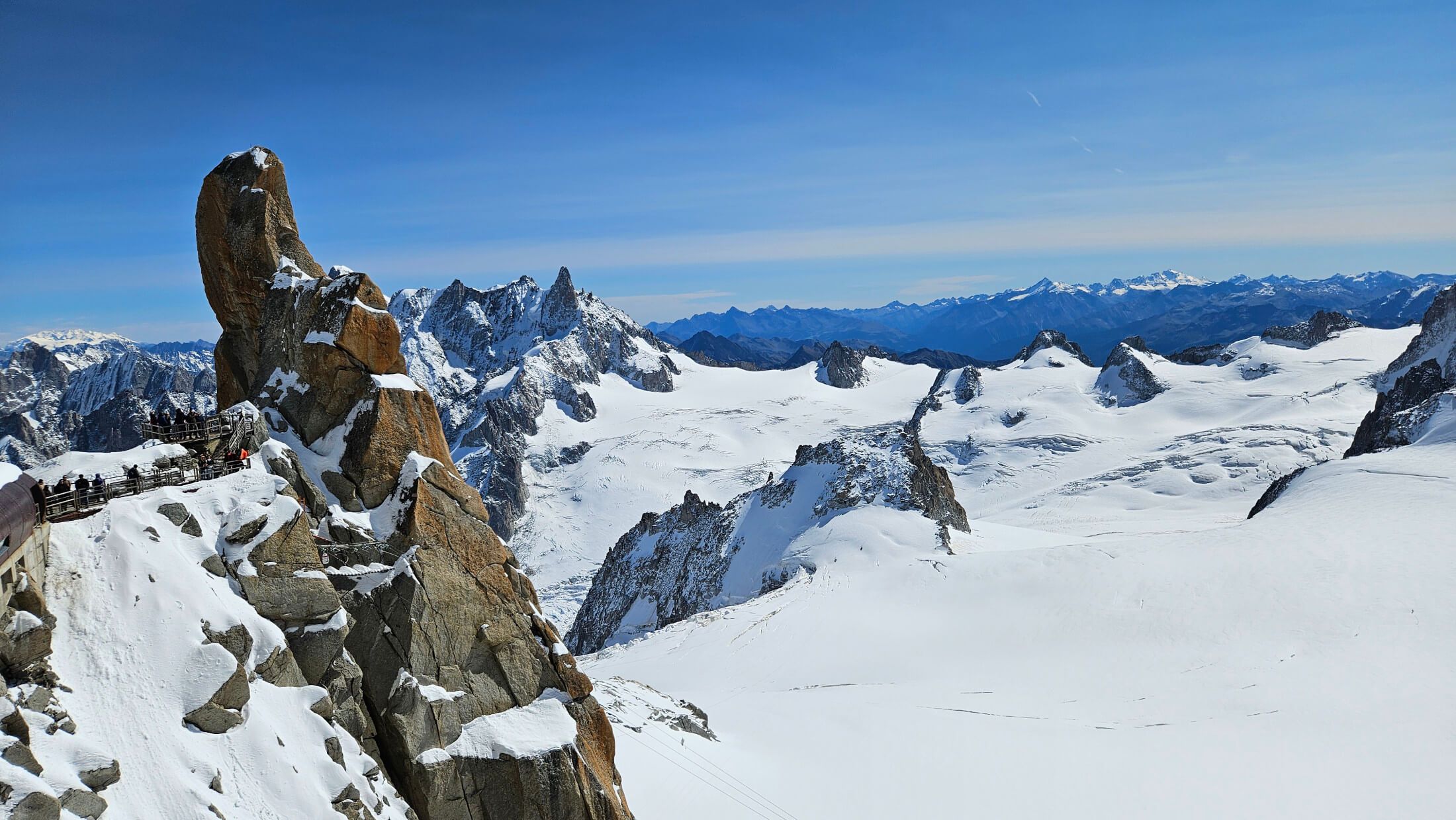 Atemberaubender Blick auf schneebedeckte Berge und einen felsigen Ausblickspunkt unter klarem blauen Himmel.