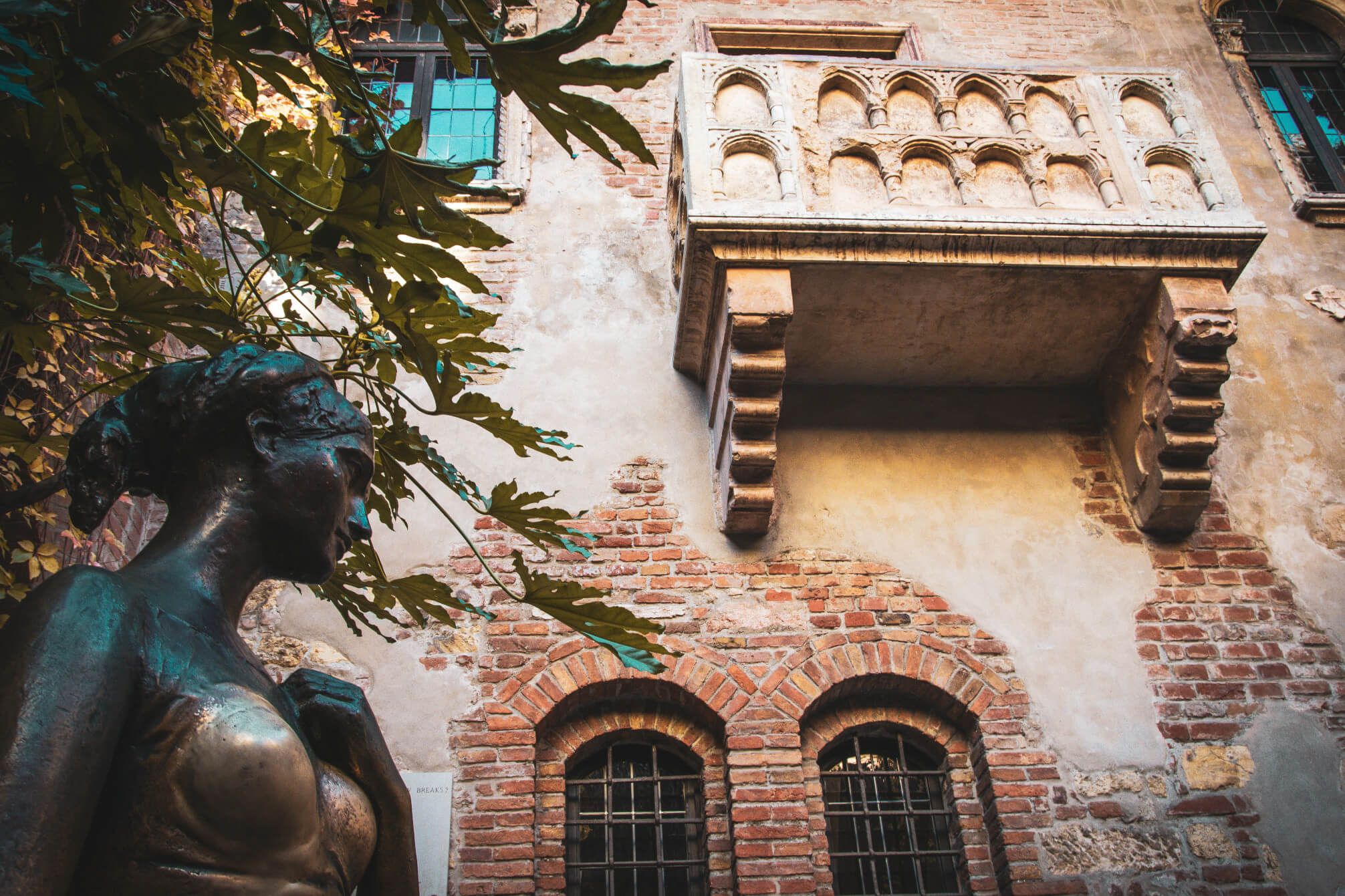 Eine Statue steht vor einer Backsteinwand mit einem Balkon und grünen Blättern, die die Szene umranden.