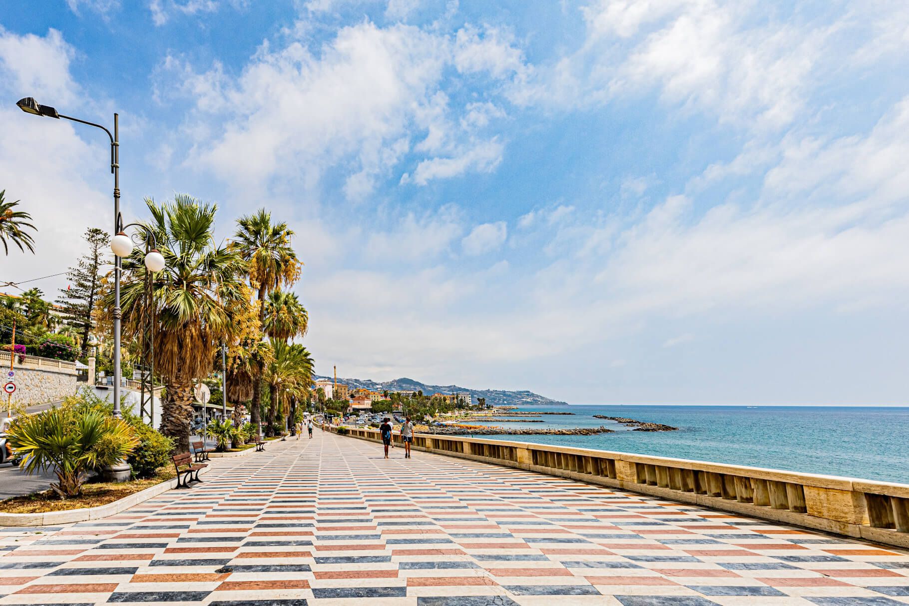 Eine schöne Strandpromenade mit Palmen, buntem Bodenmuster und Blick auf das Meer unter einem teilweise bewölkten Himmel.