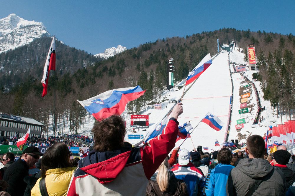 Zuschauer jubeln mit Fahnen auf einem Skihang, umgeben von schneebedeckten Bergen und einer festlichen Atmosphäre.