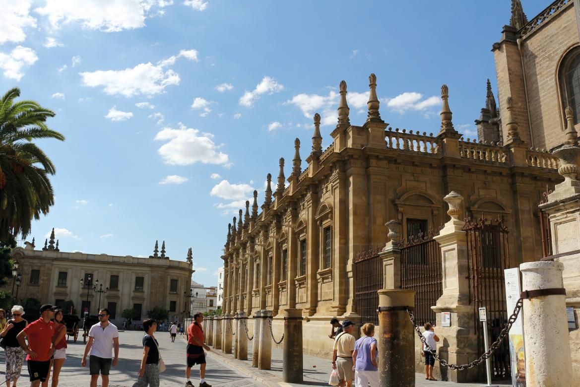 Ein sonniger Platz mit historischen Gebäuden und Spaziergängern, umgeben von Palmen und blauem Himmel.