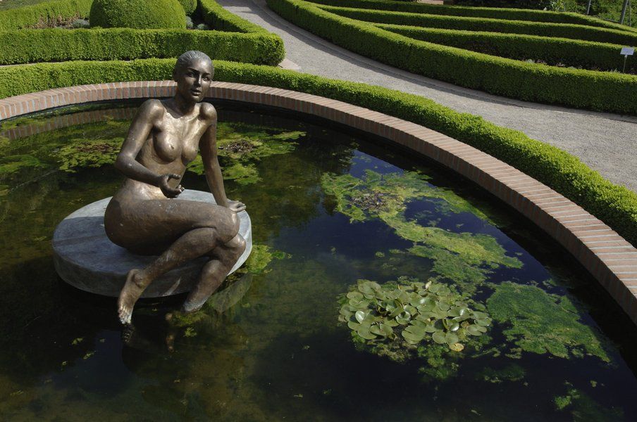 Eine nackte Statue sitzt auf einem Stein in einem Teich, umgeben von geschnittenen Hecken und Wasserpflanzen.
