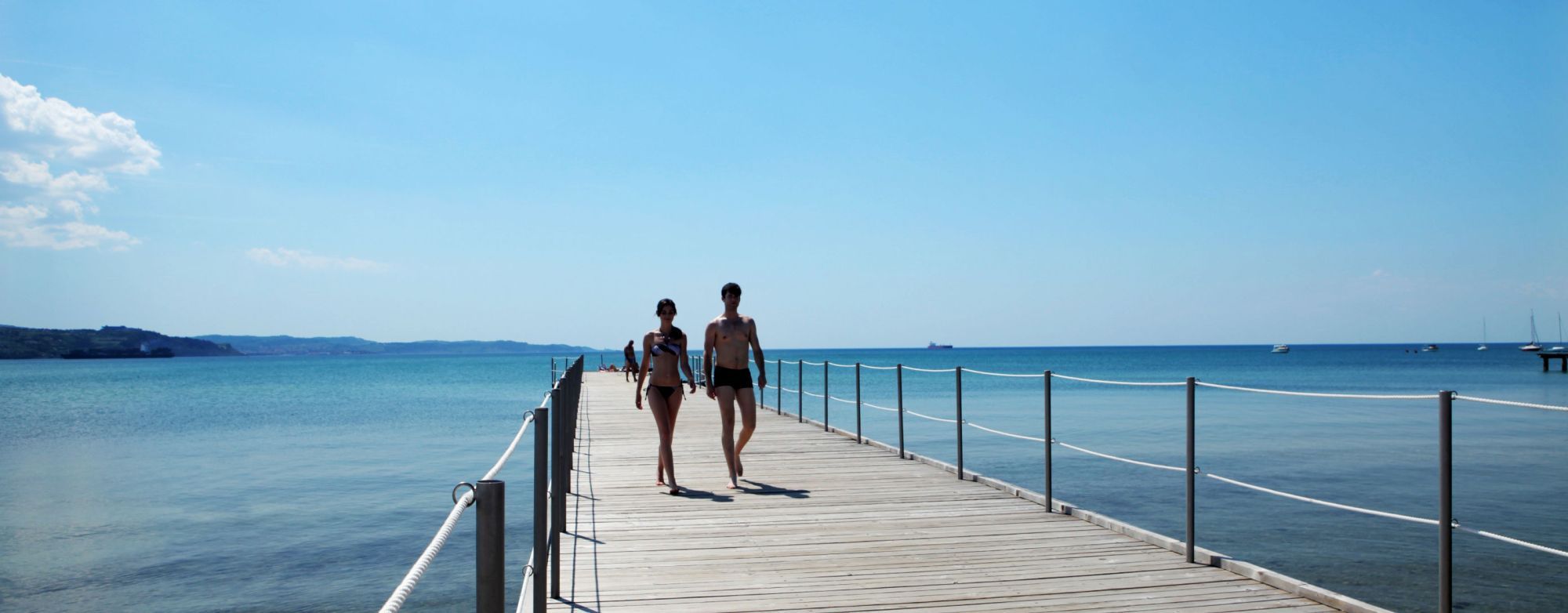 Zwei Personen spazieren auf einem Holzsteg entlang des klaren Wassers an einem sonnigen Tag.