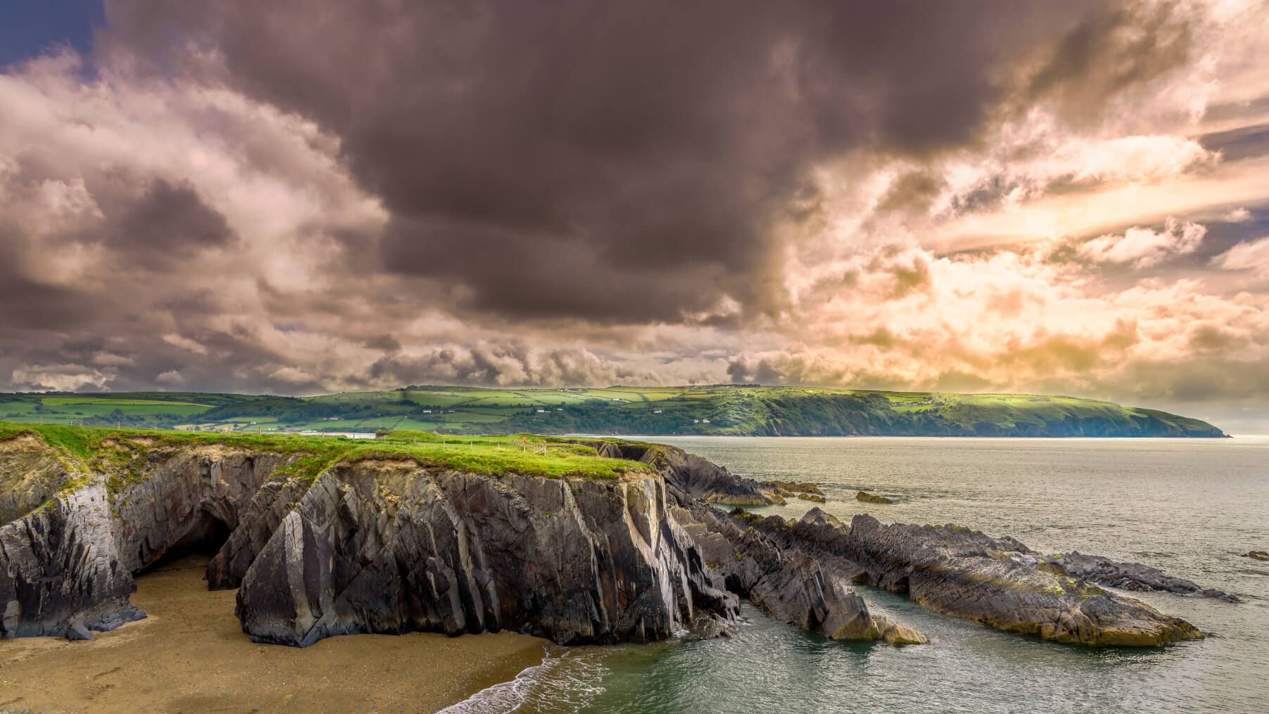 Eine dramatische Küstenlandschaft in Wales mit Felsen, grünem Hügel und bewölktem Himmel. Das Meer glitzert im Sonnenlicht.