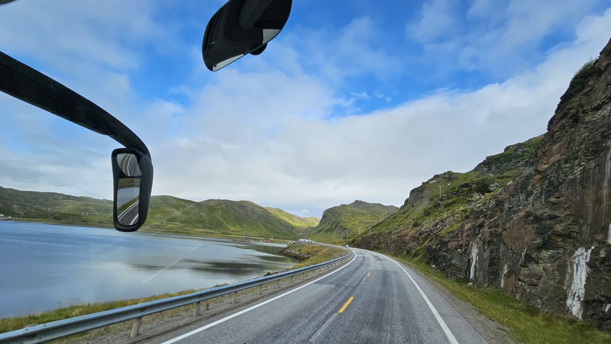 Eine kurvenreiche Straße entlang eines ruhigen Gewässers, umgeben von grünen Hügeln und dramatischer Landschaft.