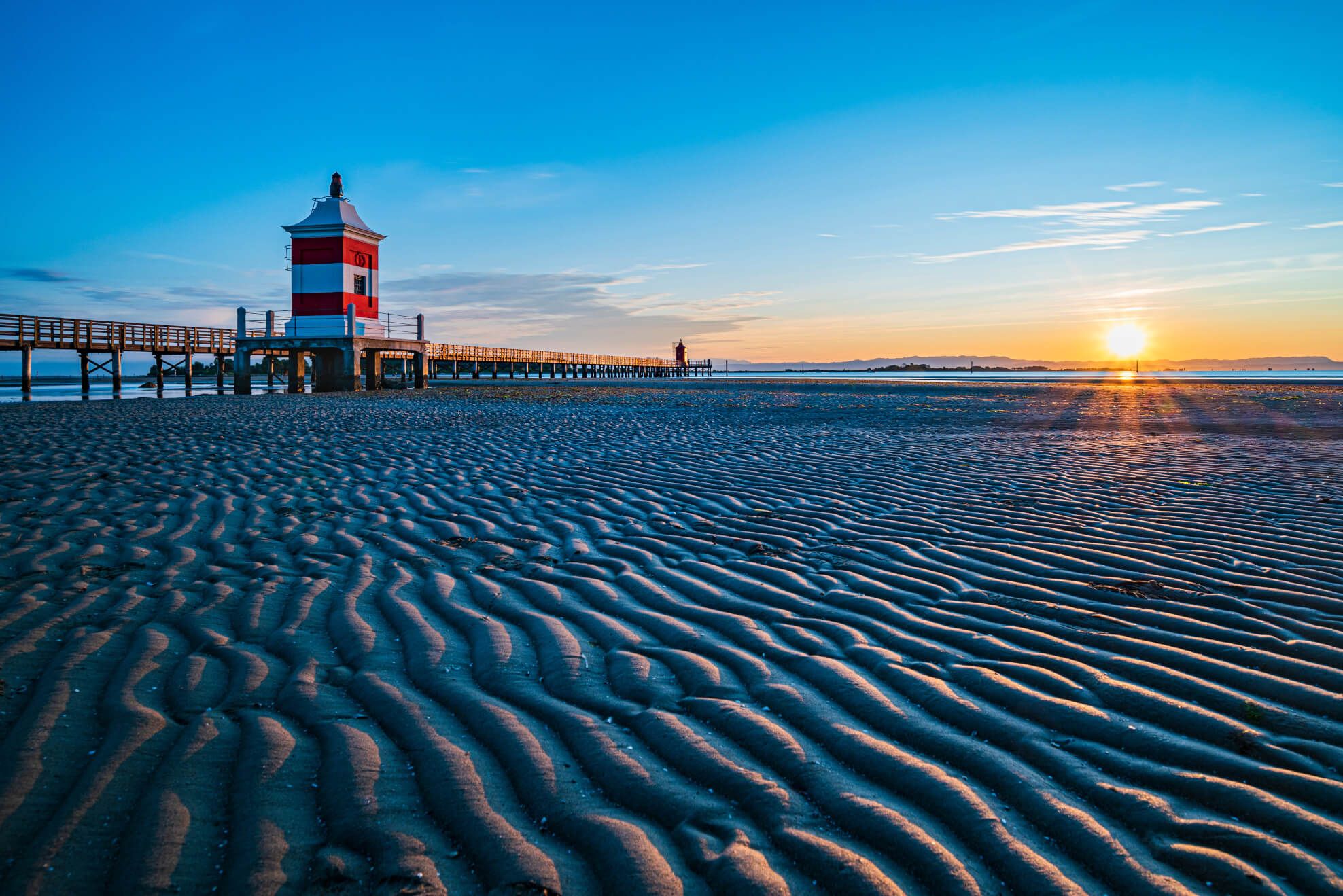 Ein schöner Strand mit einem roten Leuchtturm und einem Holzsteg, während die Sonne am Horizont aufgeht.