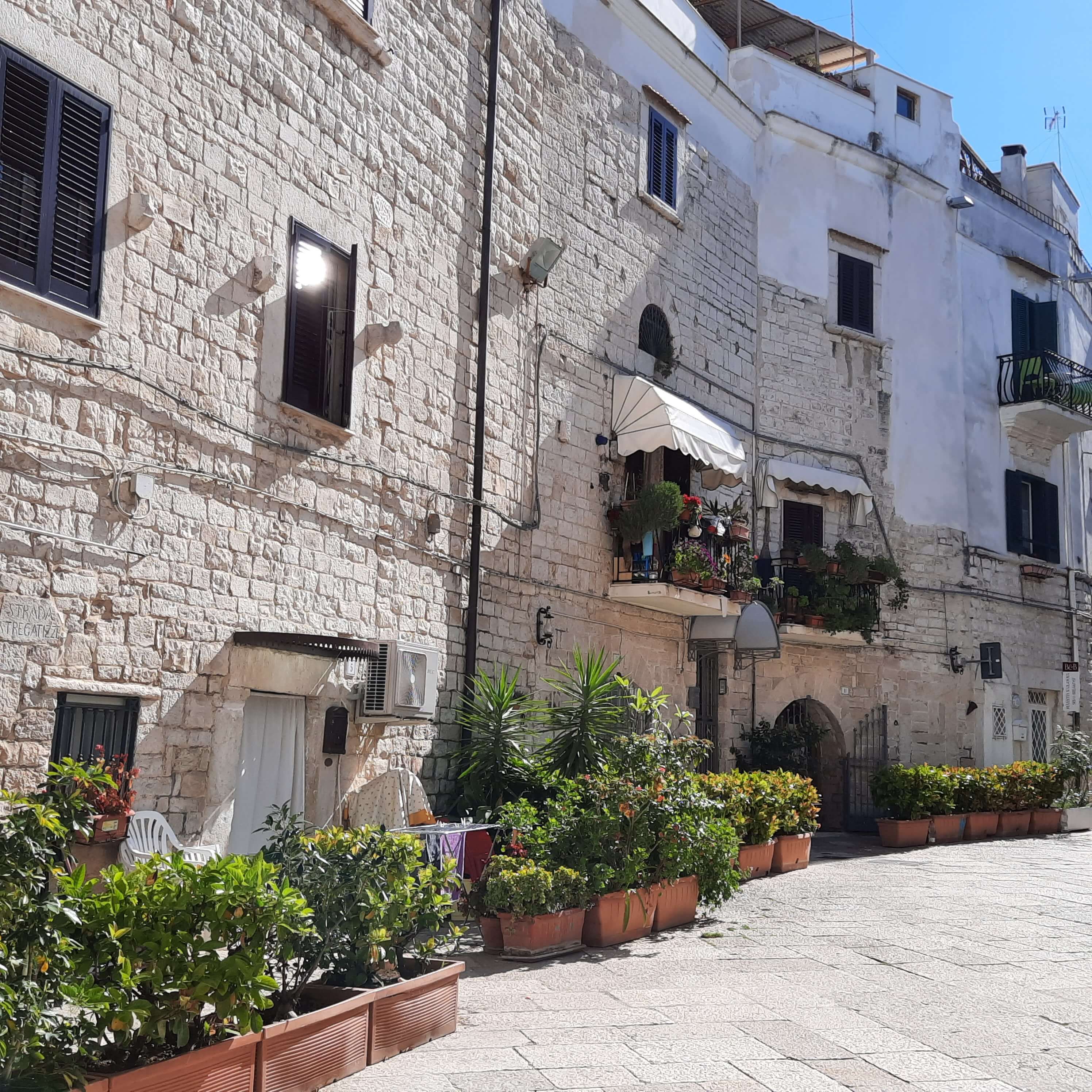 Eine sonnige Gasse mit steinernen Häusern und blühenden Pflanzen in Töpfen, ideal für einen ruhigen Spaziergang.