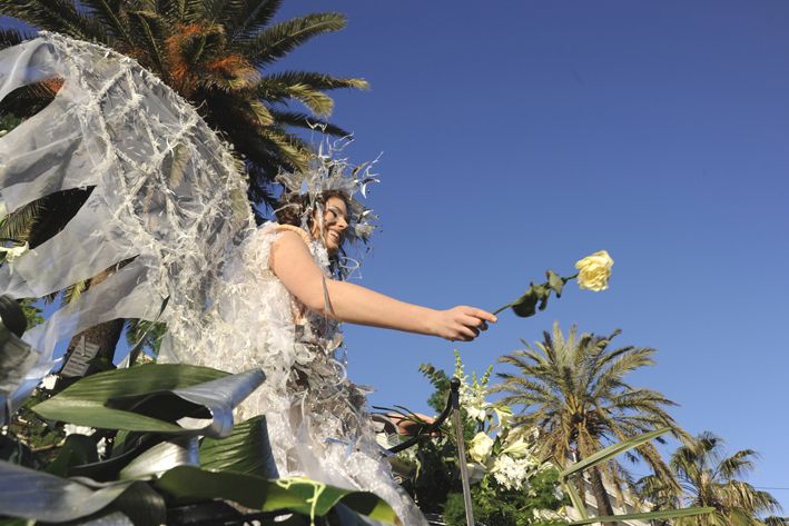 Eine Person mit Flügeln aus transparentem Material steht unter Palmen und hält eine gelbe Rose in der Hand.