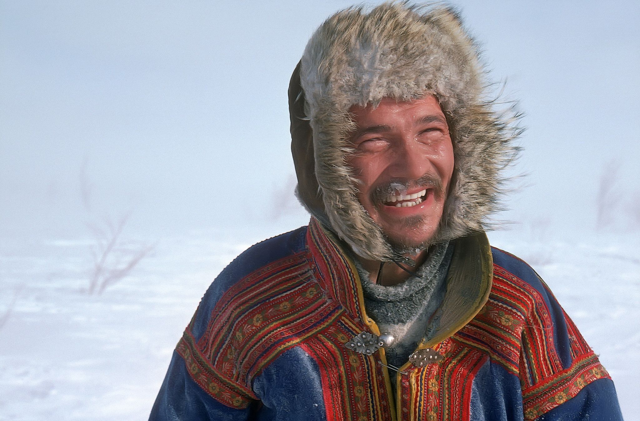 Ein lächelnder Mann in traditioneller Kleidung steht im Schnee, umgeben von frostiger Landschaft und Nebel.
