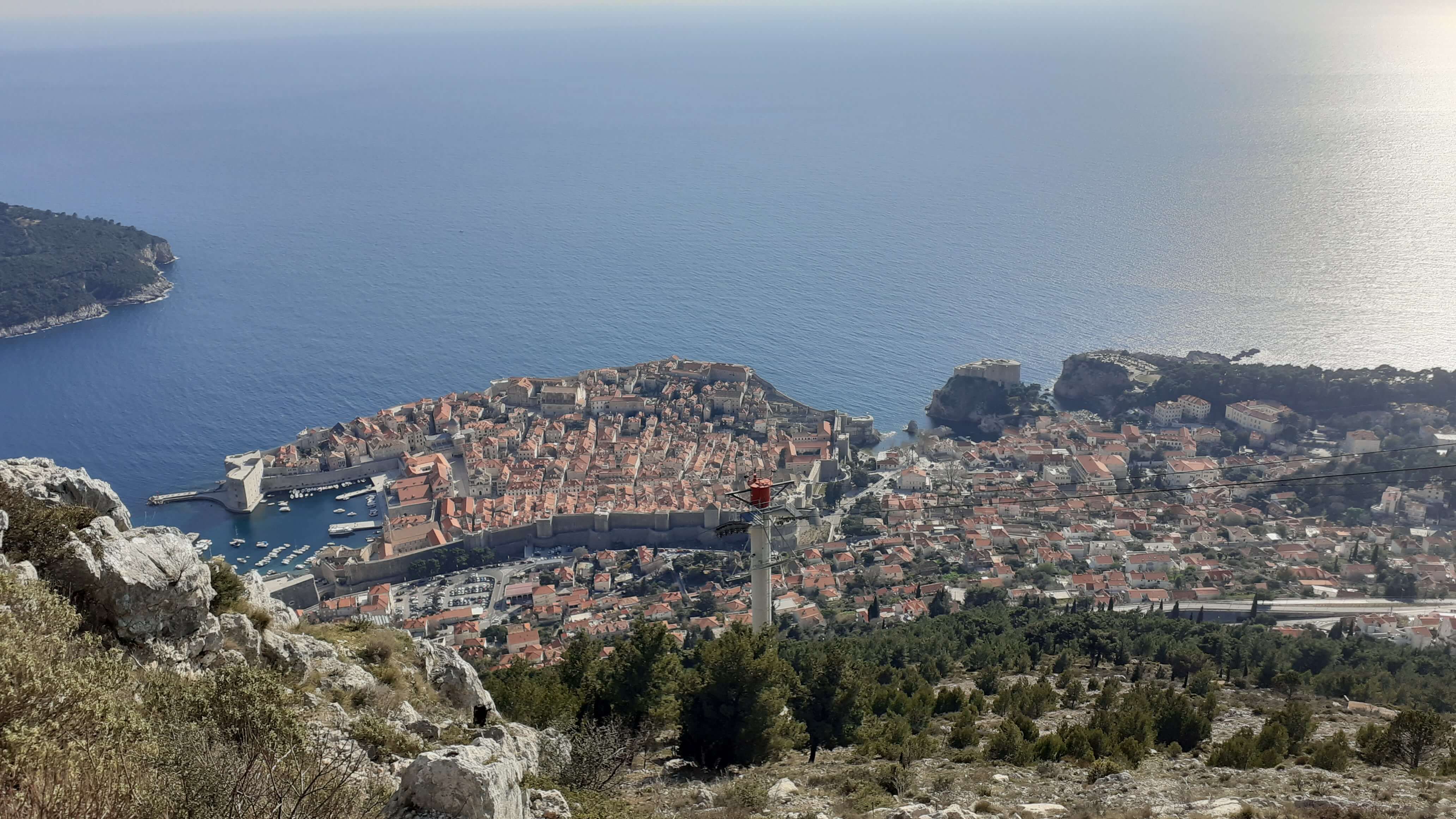 Blick von oben auf die Küstenstadt Dubrovnik mit dem tiefblauen Meer im Hintergrund und grünen Hügeln.