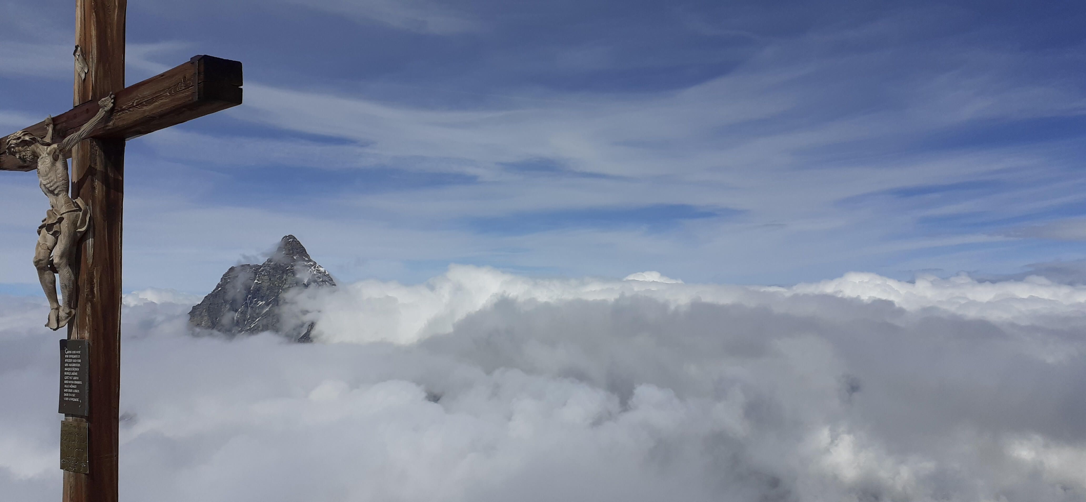 Ein Kreuz mit Jesusfigur über schneebedeckten Bergen, umgeben von Wolken und blauem Himmel. Atemberaubende Aussicht!