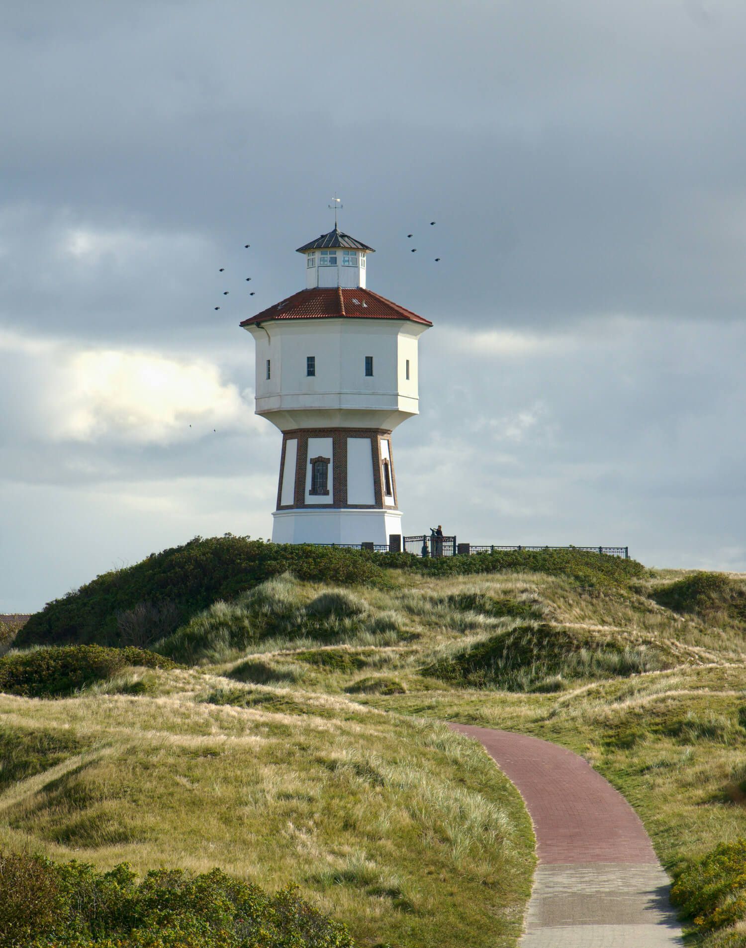 Ein Wasserturm auf einer grünen Düne, umgeben von grasbewachsenen Hügeln und einem Weg, wolkiger Himmel. (C) Foto: K. Rolf