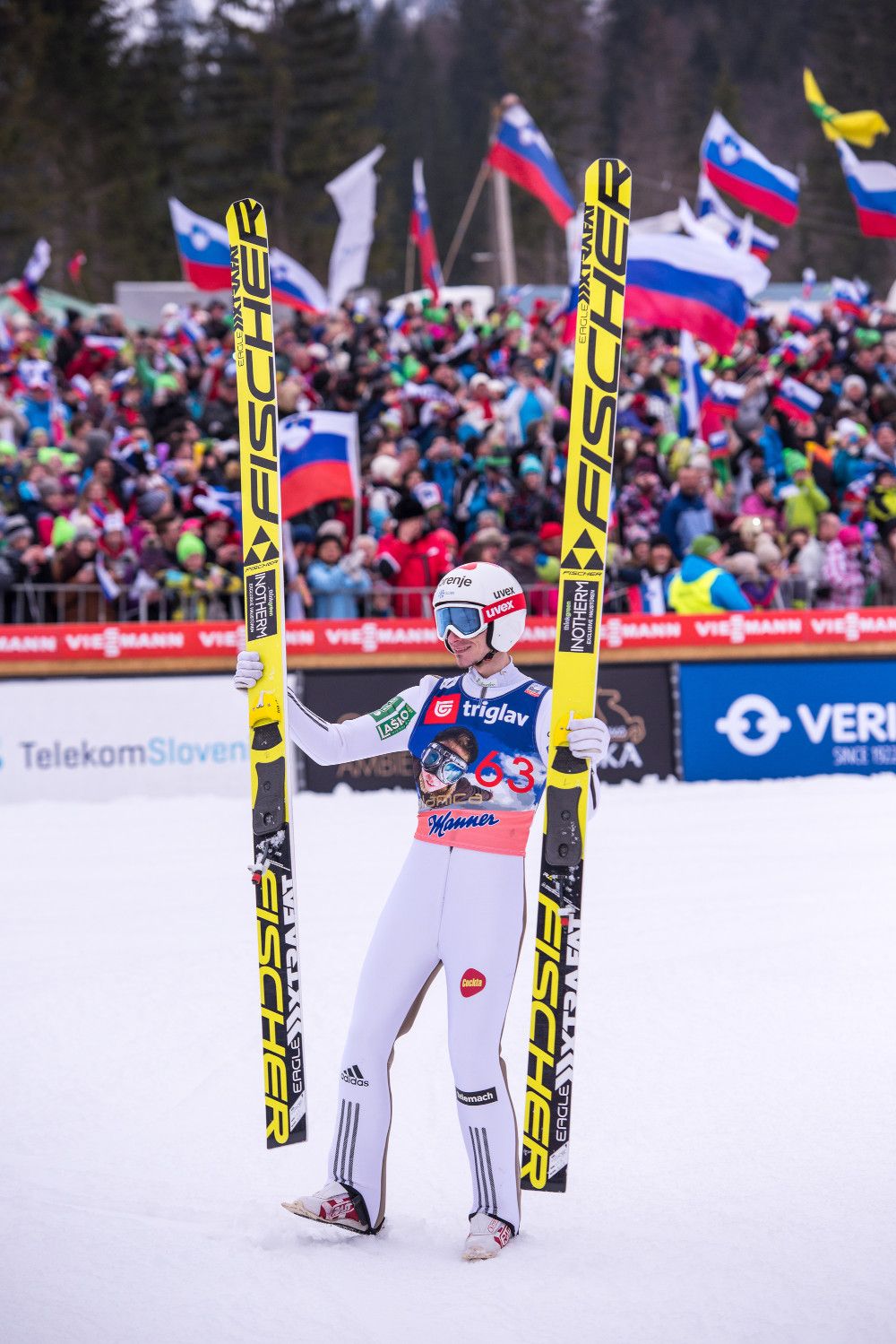 Ein Skispringer in weißem Anzug feiert vor einer jubelnden Menge mit Fahnen im Hintergrund.