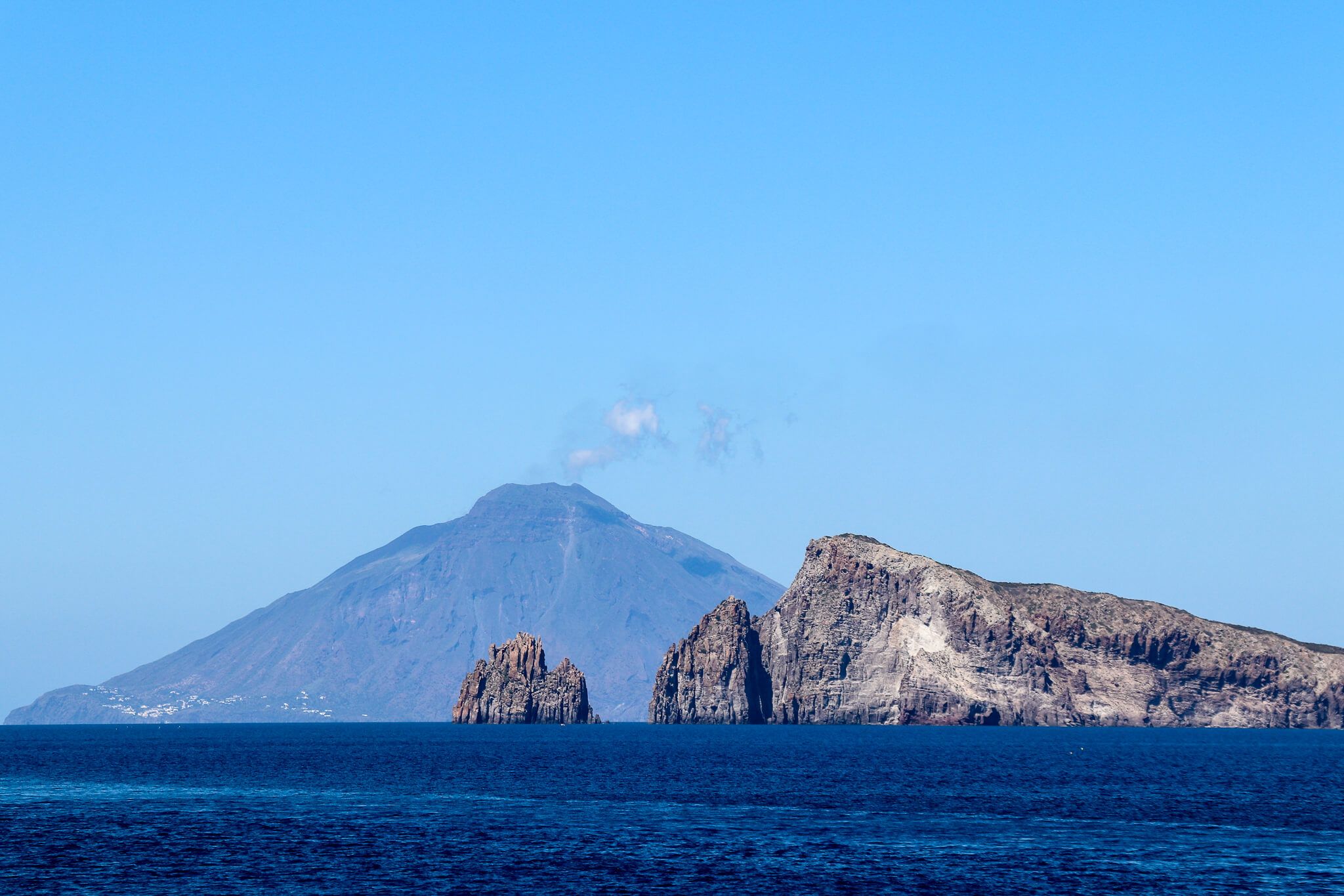 Vulkanlandschaft mit Meer und zwei Felsen im Vordergrund, blauer Himmel ohne Wolken. Sehr friedliche Szenerie.