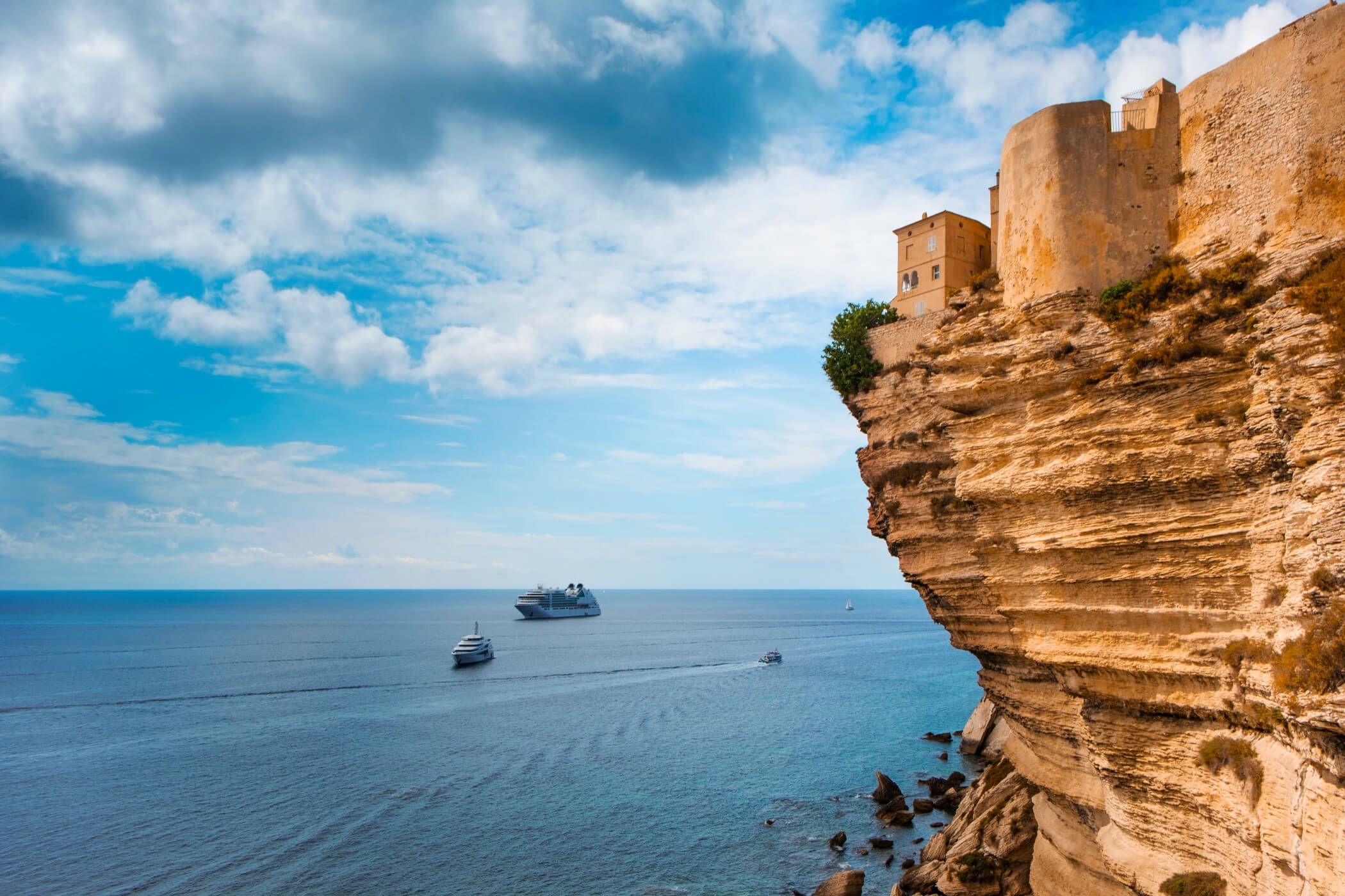 Eine Felsenküste mit einem Gebäude oben, darunter Boote und ein weites Meer unter einem klaren Himmel.