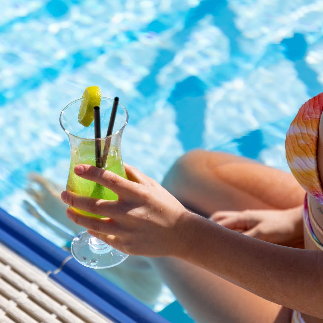 Eine Person sitzt am Poolrand und hält ein Getränk mit Limettenscheibe und zwei Strohhalmen in der Hand.