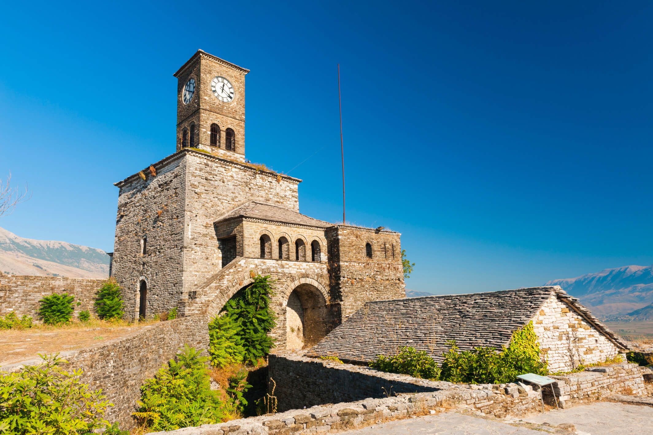 Eine alte steinerne Uhrturm-Burg im klaren blauen Himmel, umgeben von grüner Landschaft und Bergen.