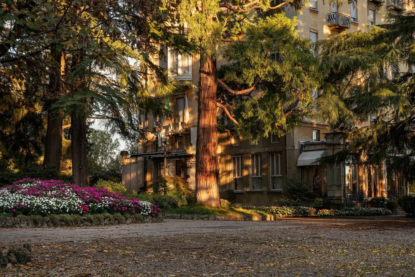Ein Garten mit bunten Blumen und hohen Bäumen vor einem historisches Gebäude im warmen Licht der Abendsonne.