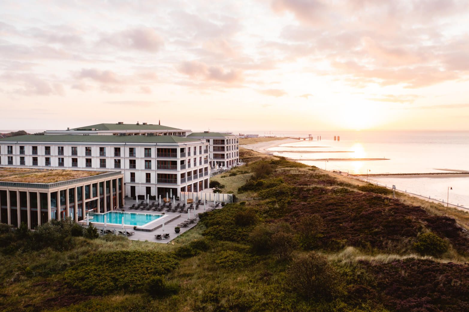 Moderne Hotelanlage mit Pool am Meer, umgeben von Natur, bei Sonnenuntergang. Ruhe und Entspannung garantiert.