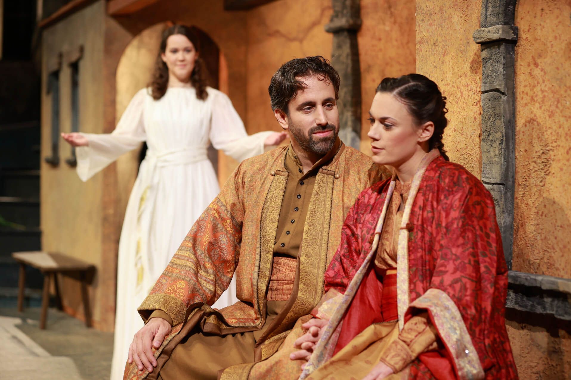 Drei Schauspieler auf der Bühne: ein Mann und eine Frau im Gespräch, eine dritte Person im Hintergrund.