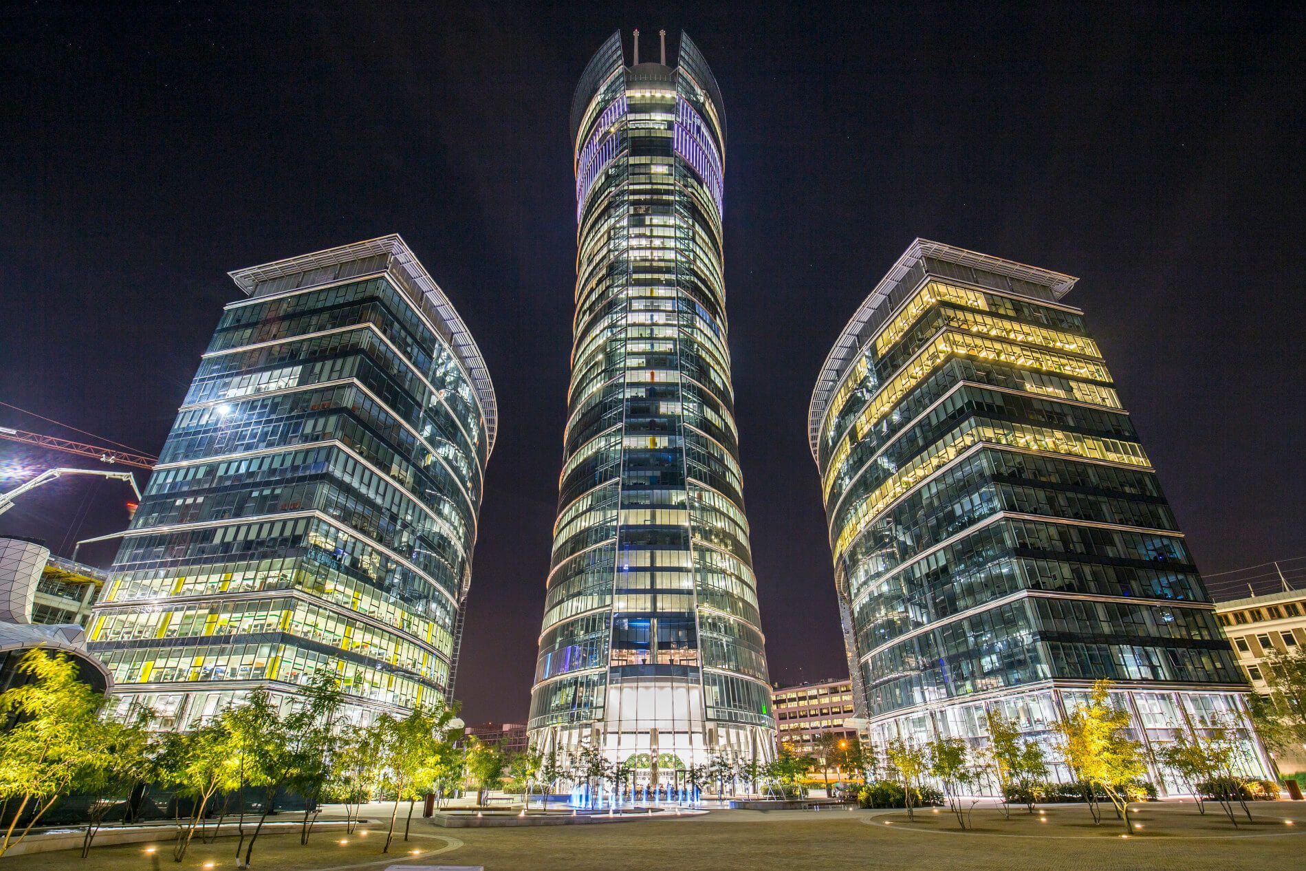 Moderne Wolkenkratzer leuchten nachts, umgeben von Bäumen und einem klaren Himmel. Ein beeindruckendes Stadtbild.