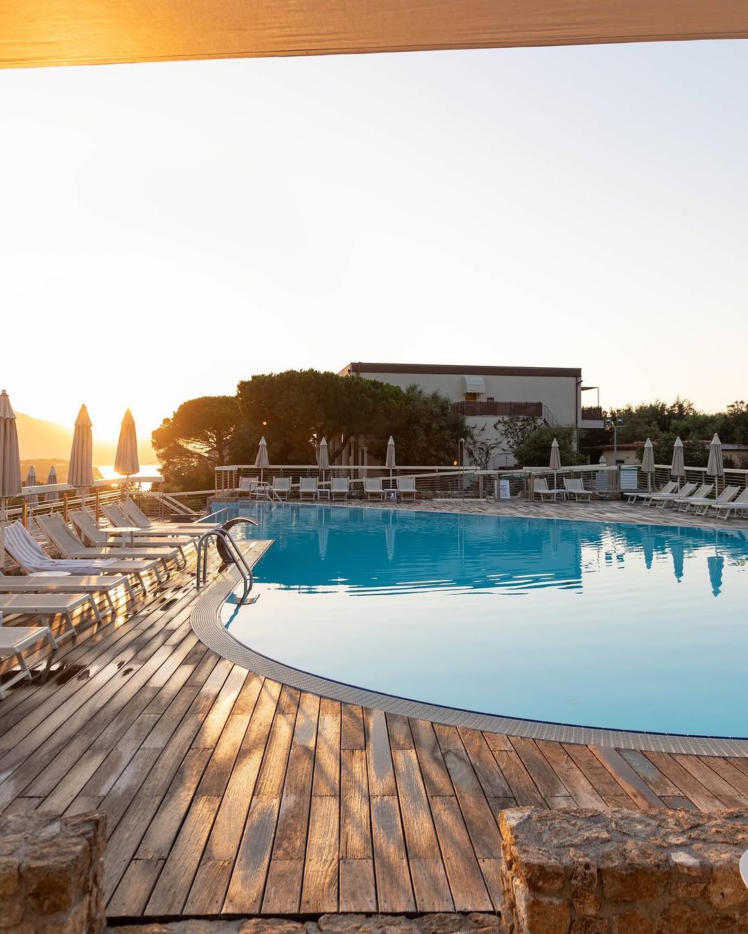 Sonnenuntergang über einem Pool mit Liegen und Sonnenschirmen, umgeben von Bäumen und einem ruhigen Ambiente.