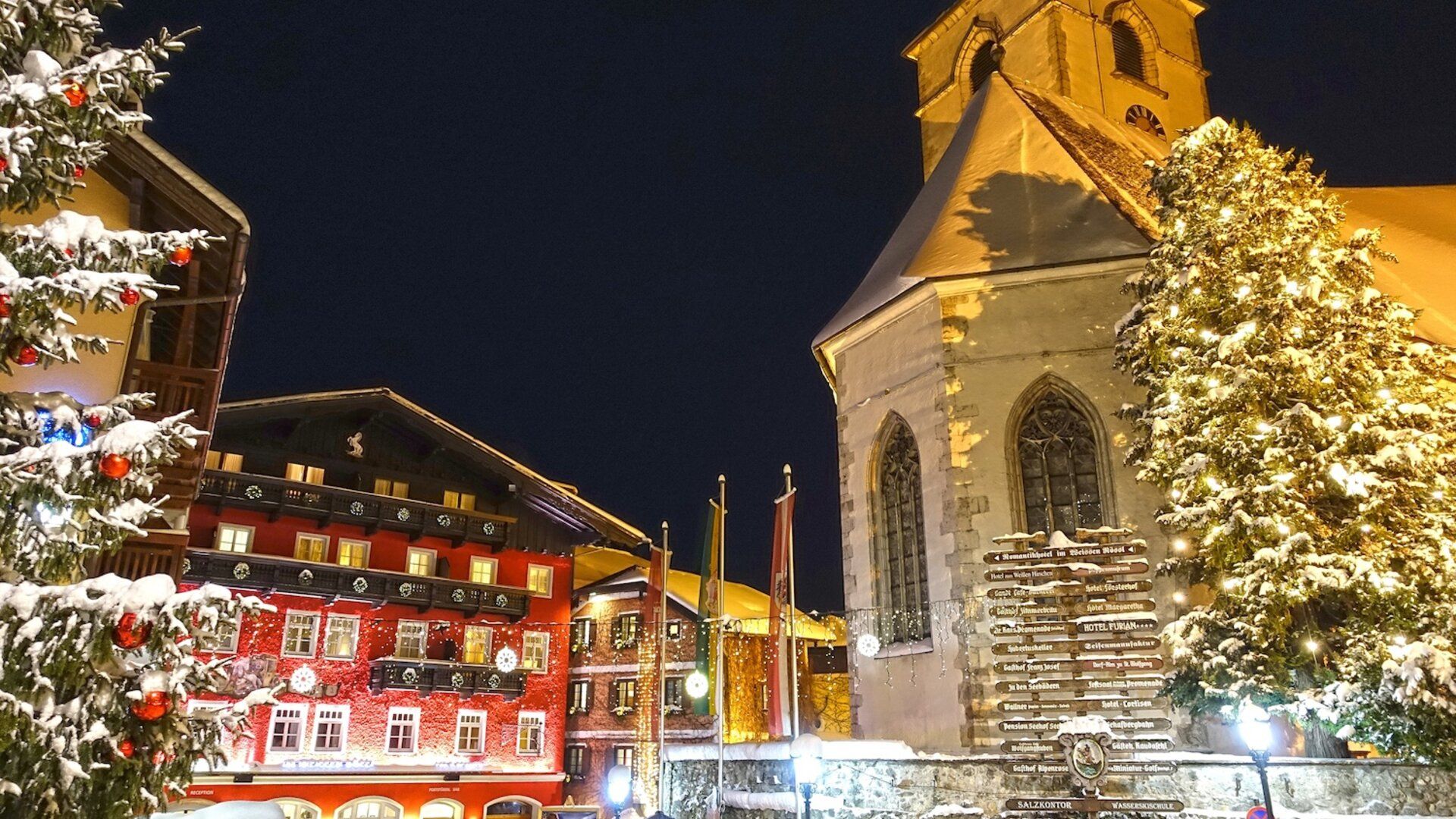 Eine verschneite Szene bei Nacht mit festlich beleuchteten Gebäuden und einem Weihnachtsbaum in einem Dorf.