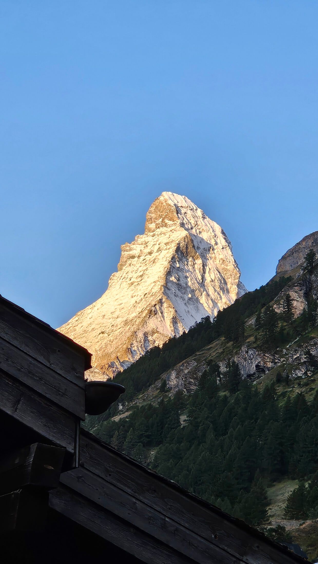 Ein hoch aufragender Berggipfel wird von Sonnenlicht erleuchtet, vor blauem Himmel und bewaldeten Hängen.