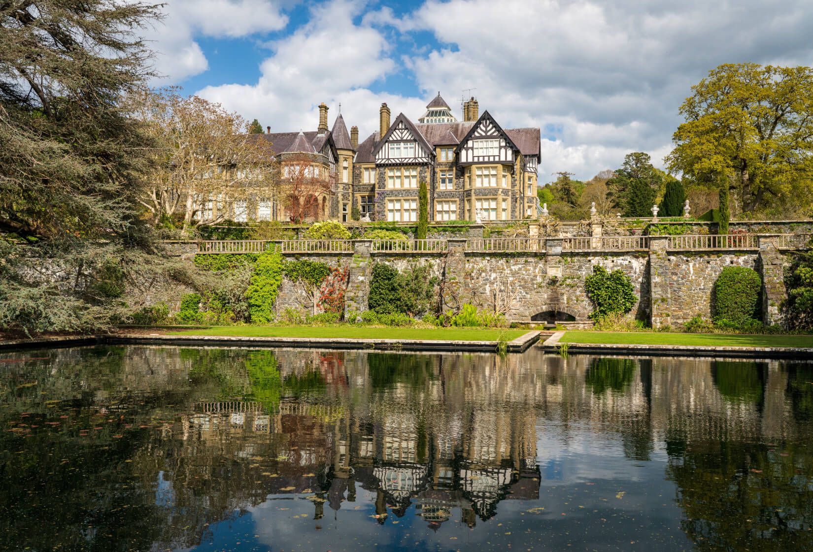 Ein elegantes Herrenhaus spiegelt sich im ruhigen Wasser eines Teichs, umgeben von üppigem Grün und Bäumen.