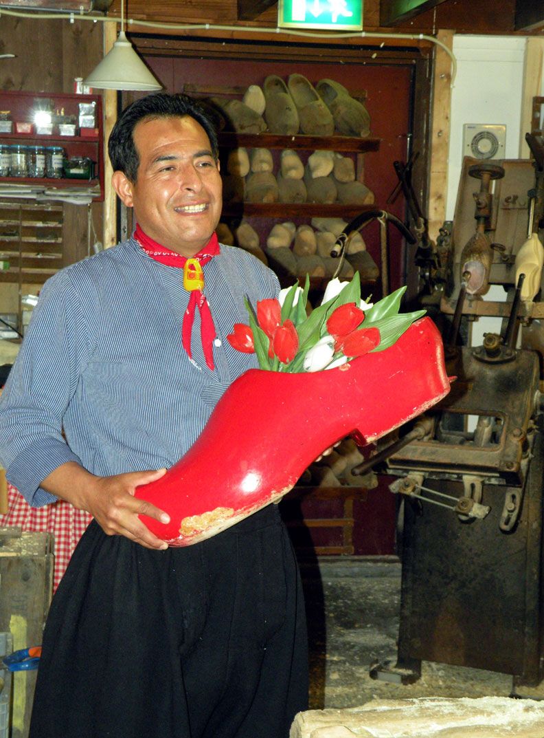 Ein Mann hält einen roten Holzschuh mit Blumen in der Hand, lächelt in einer Werkstatt voller Schuhe.