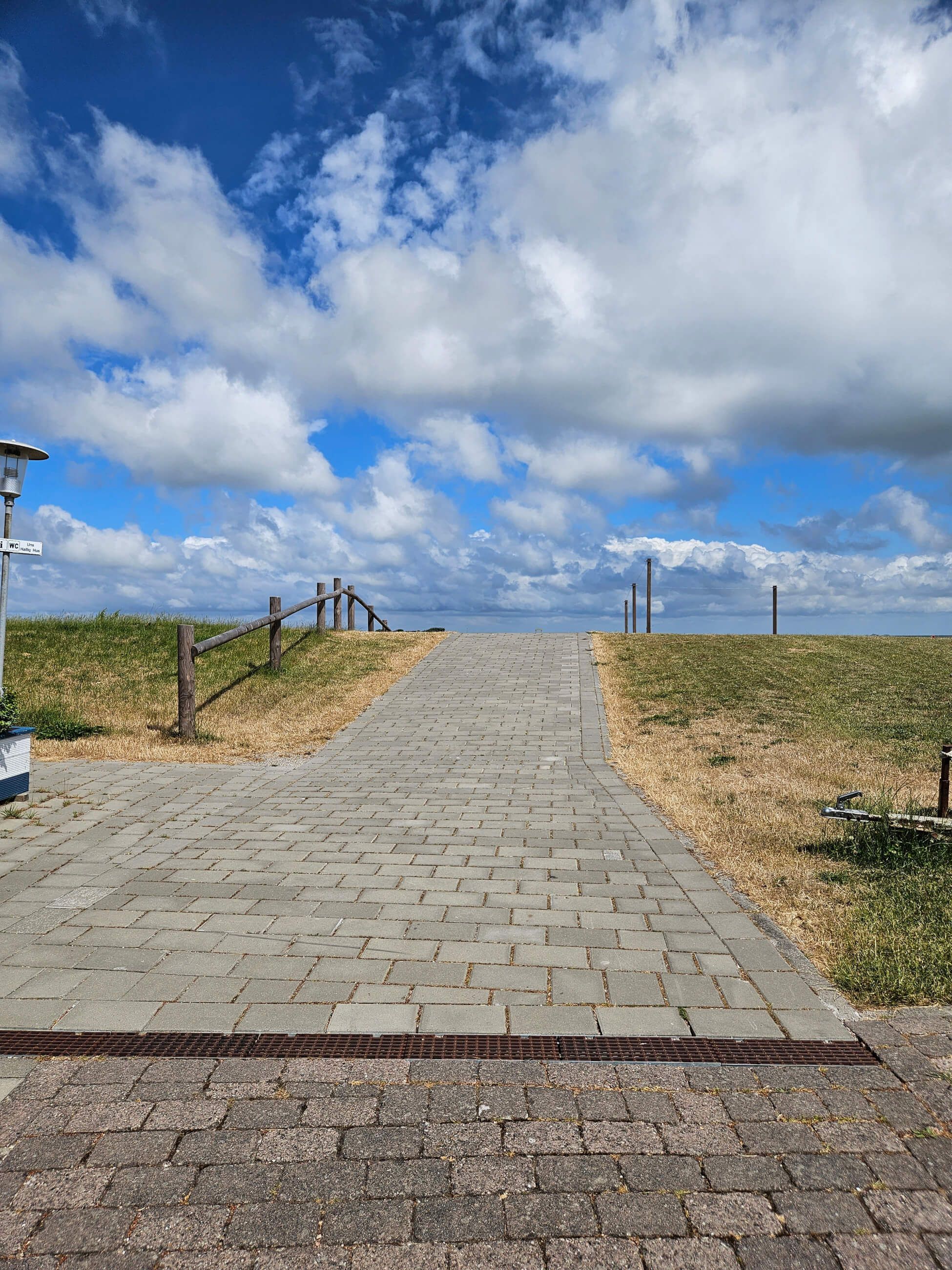 Ein gepflasterter Weg führt zu einer grünen Fläche unter einem blauen Himmel mit Wolken. Ruhe und Weite ausstrahlend.