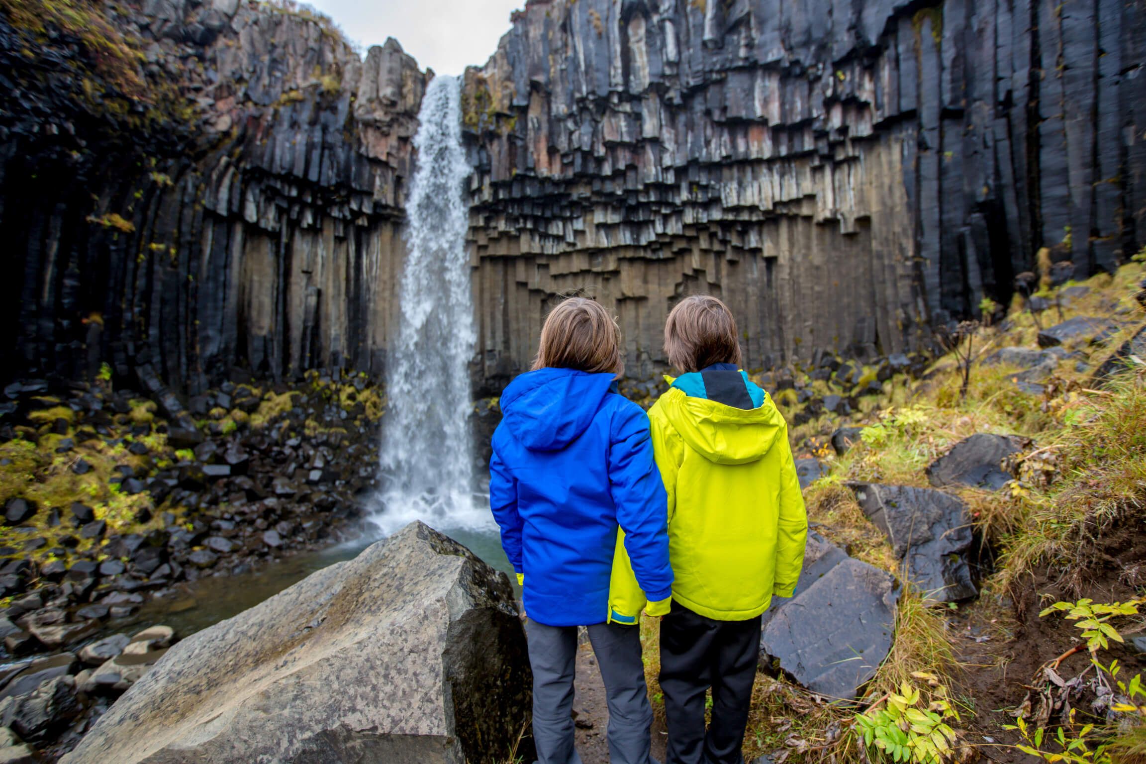Zwei Kinder stehen vor einem Wasserfall, umgeben von beeindruckenden Felsen und herbstlicher Natur.