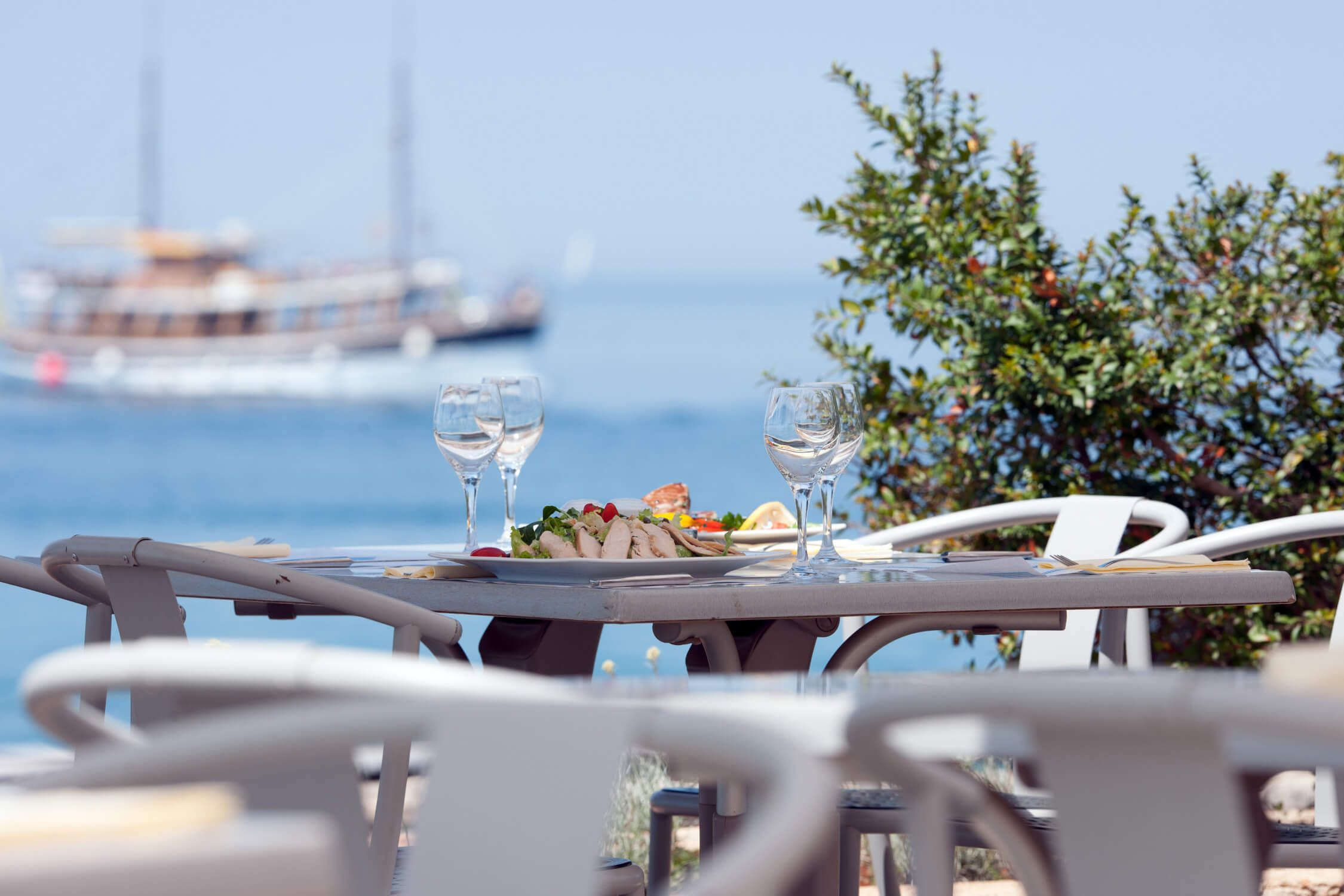 Ein gedeckter Tisch mit Essen und Gläsern, Blick auf das Wasser und ein Schiff im Hintergrund. Entspannte Atmosphäre.