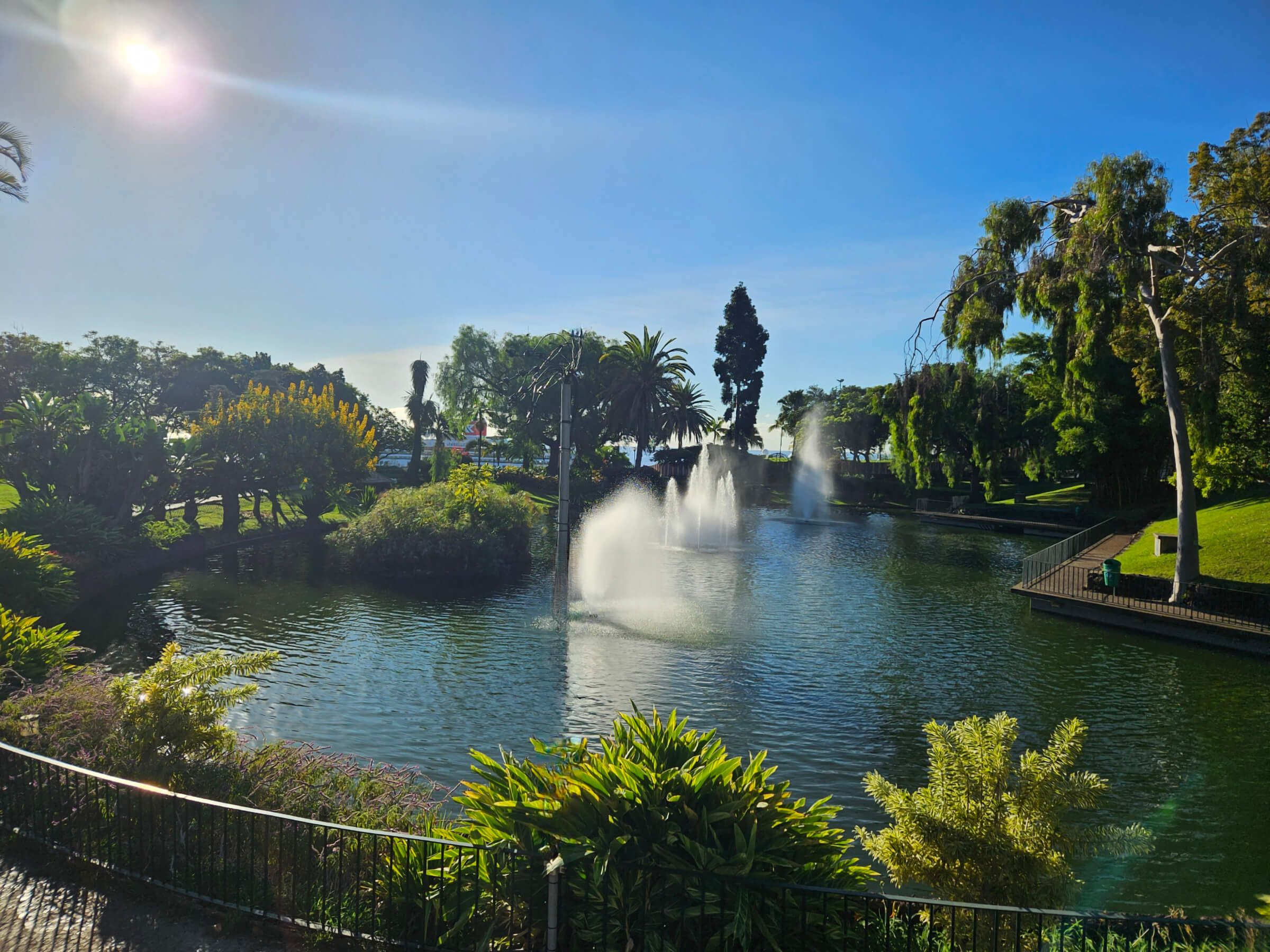 Ein ruhiger Park mit einem Teich, Wasserfontänen, grünen Bäumen und klarem Himmel bei Sonnenschein.