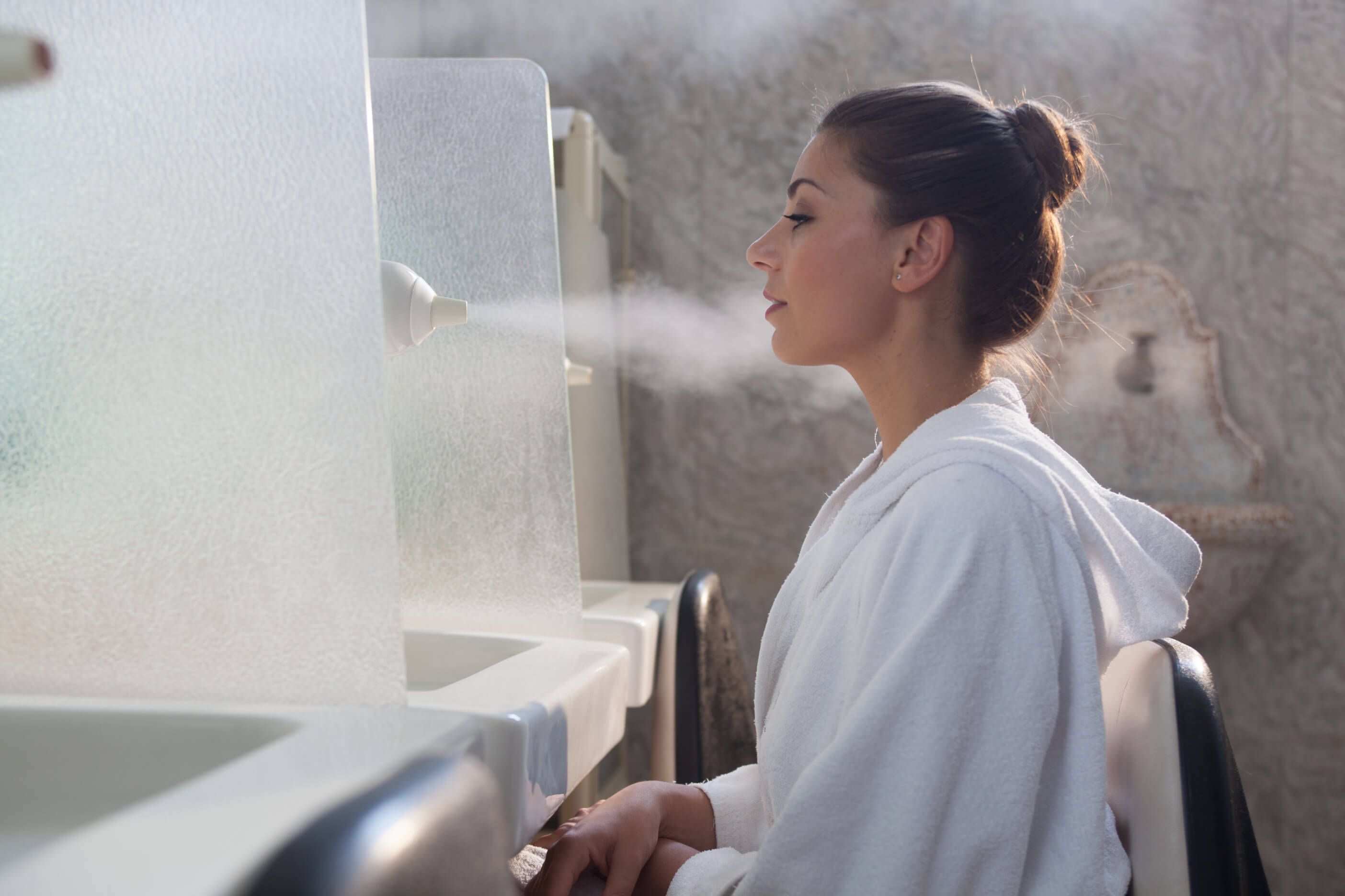 Eine Frau in einem weißen Bademantel sitzt an einem Dampfbad, während Dampf aus einer Vorrichtung strömt.