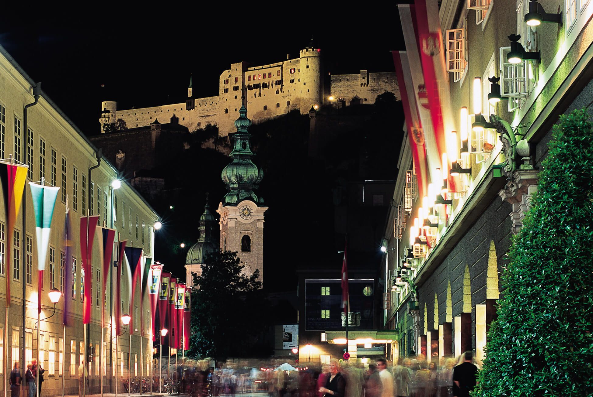 Salzburg bei Nacht, mit der Festung im Hintergrund und bunten Fahnen entlang der Straße, lebhafte Atmosphäre.