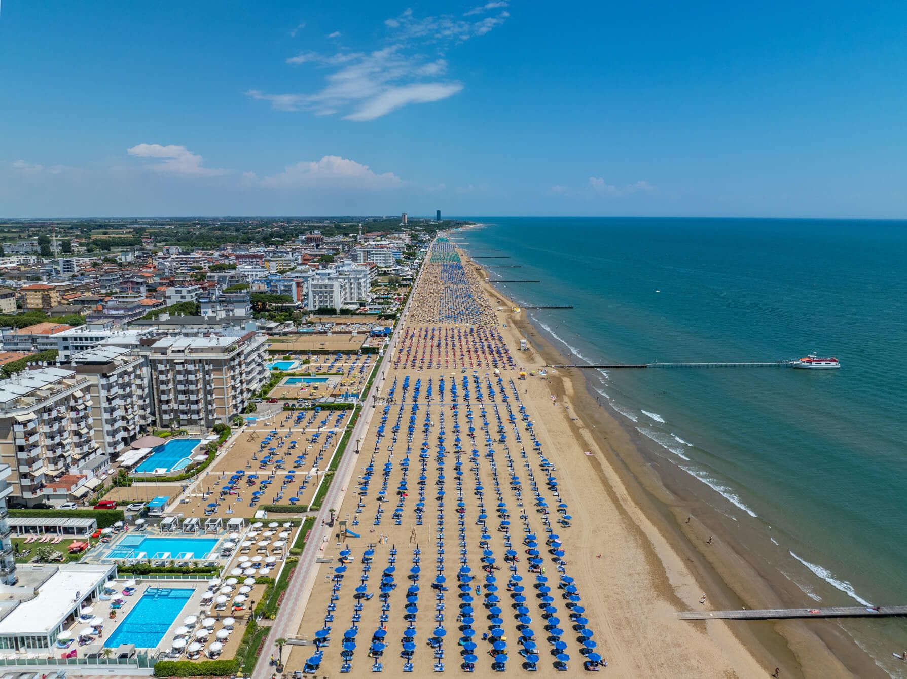 Ein weitläufiger Strand mit vielen blauen Sonnenschirmen, Hotels in der Nähe und klarem, blauem Wasser.