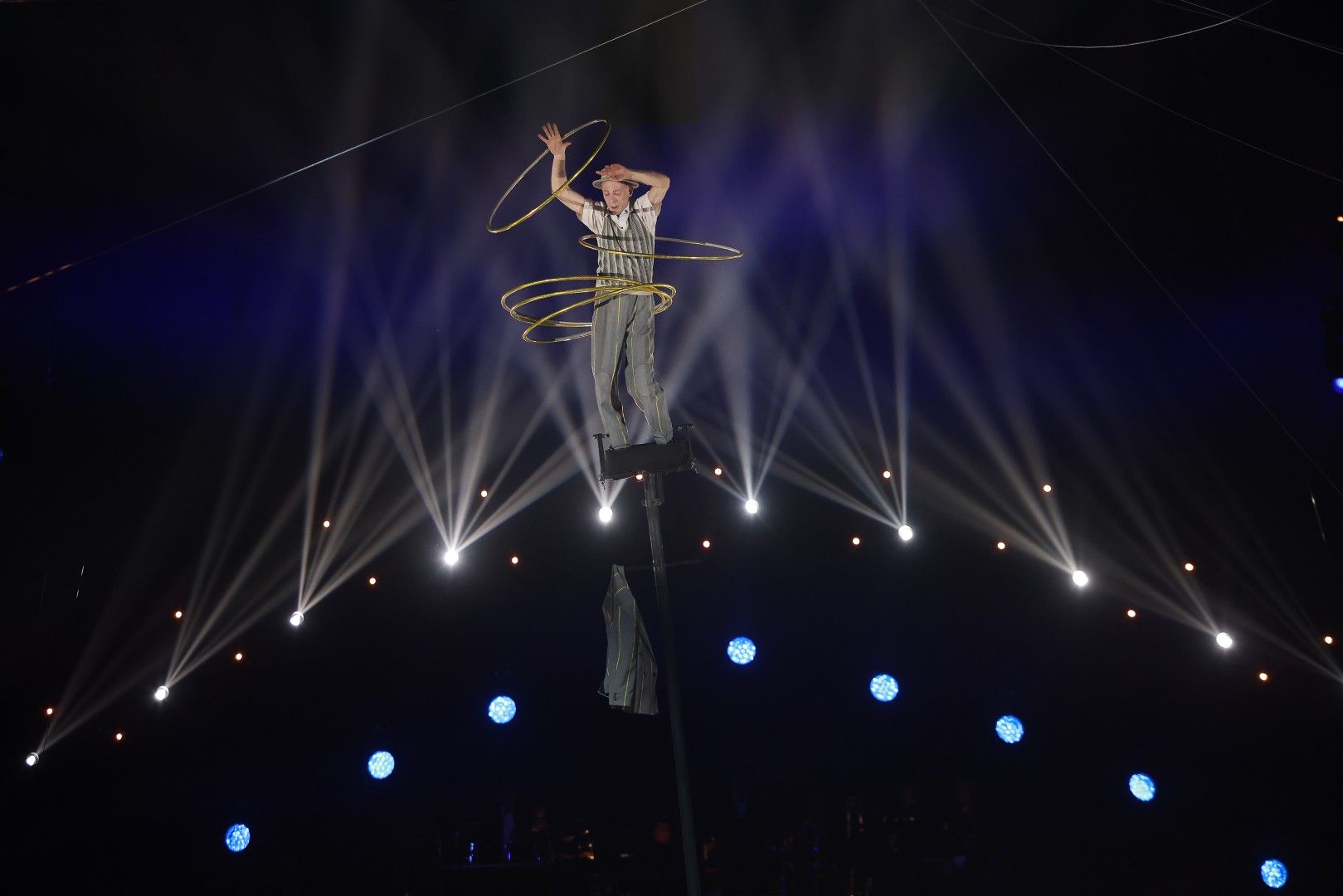Ein Akrobat steht auf einem hohen Pfahl und jongliert mit Reifen, umgeben von bunten Lichtern und einer spannenden Atmosphäre.